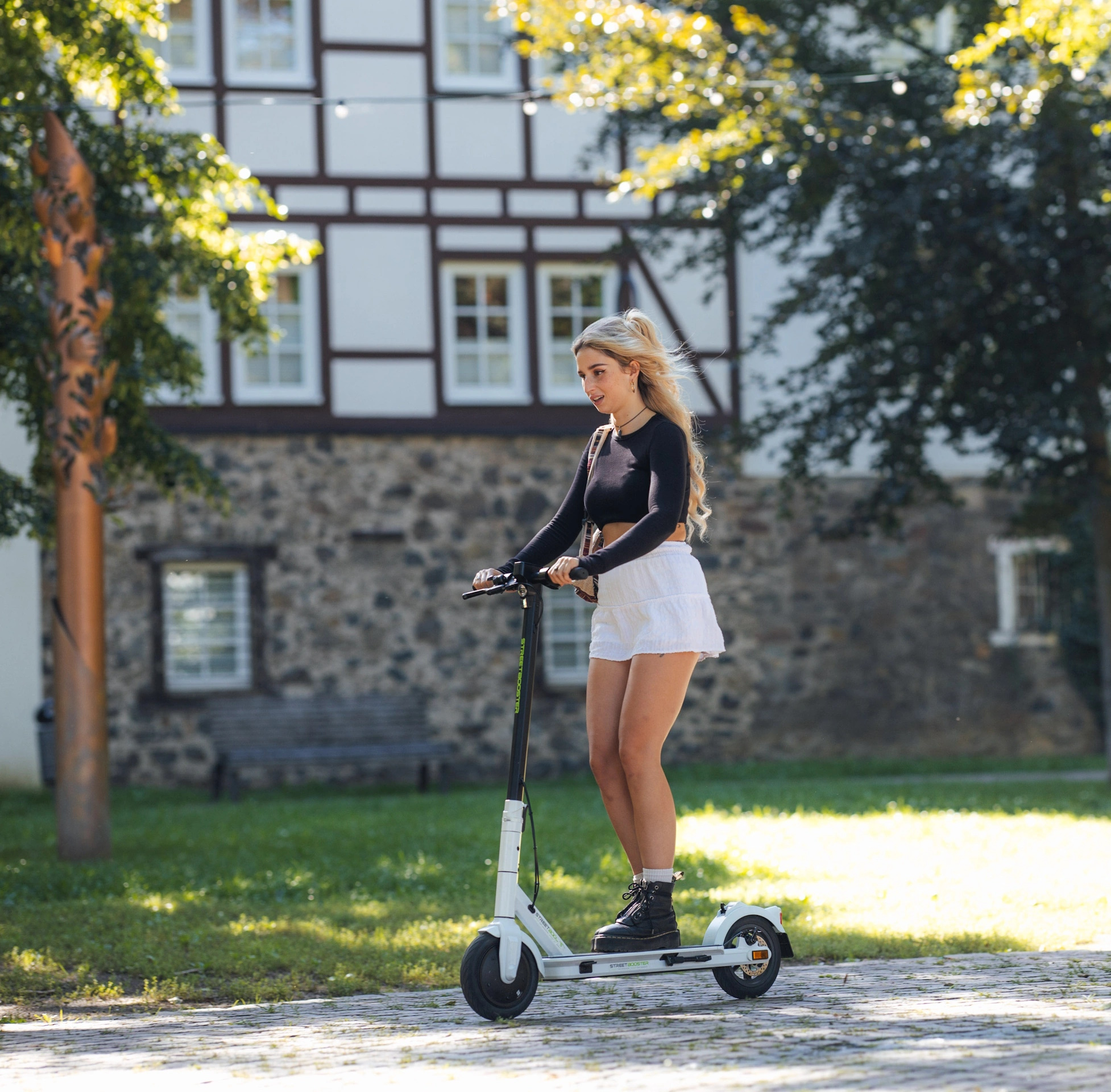 Blonde Frau auf einem Roller, trägt ein schwarzes Top, einen weißen Rock und Stiefel. Hintergrund ist ein Gebäude und Bäume.