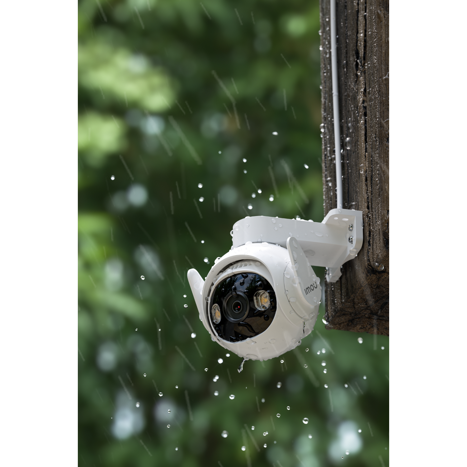 Biała kamera bezpieczeństwa na drewnianym słupie w deszczu, z kroplami wody i rozmytym zielonym tłem.
