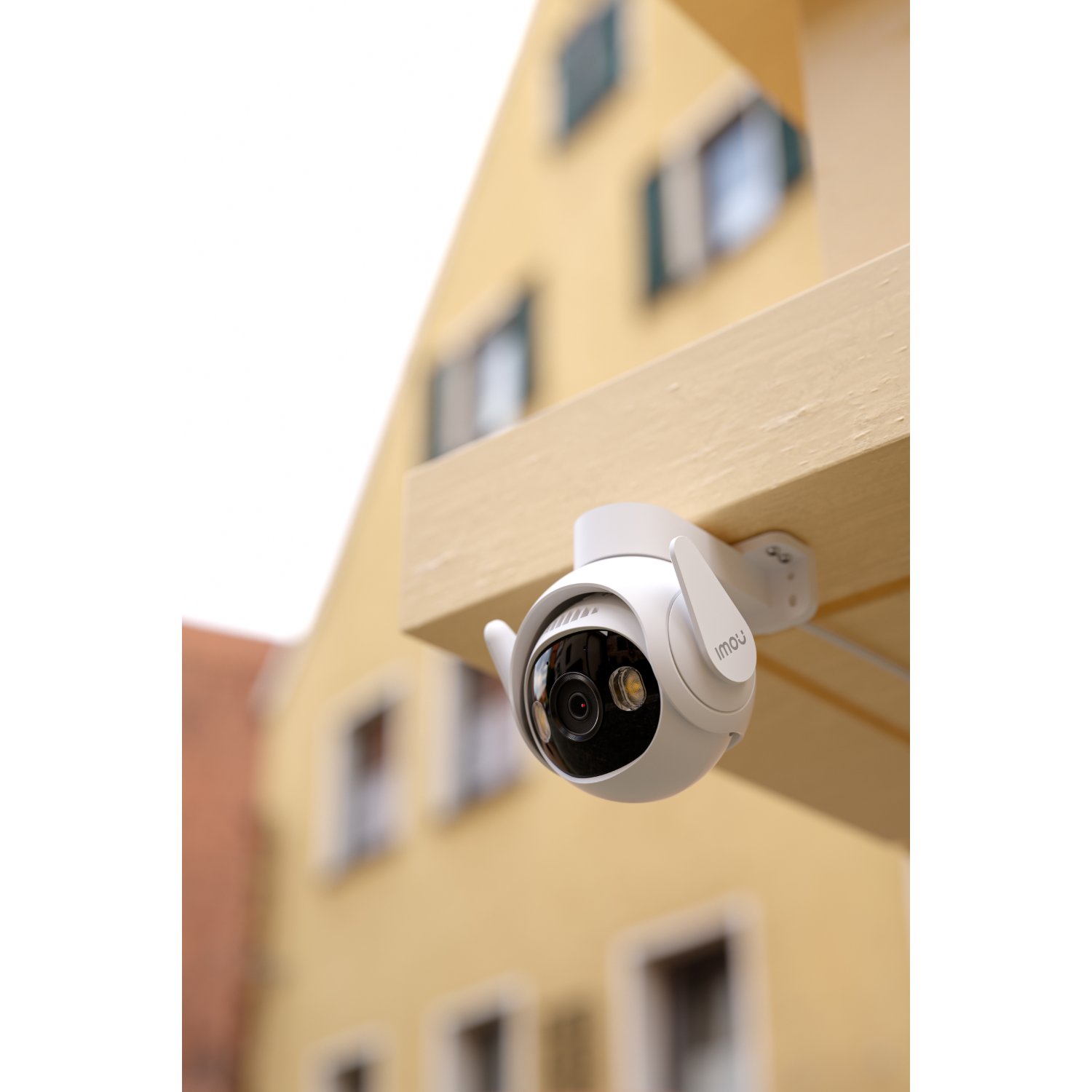 Biała kamera bezpieczeństwa zamontowana na drewnianej belce; żółty budynek w tle.