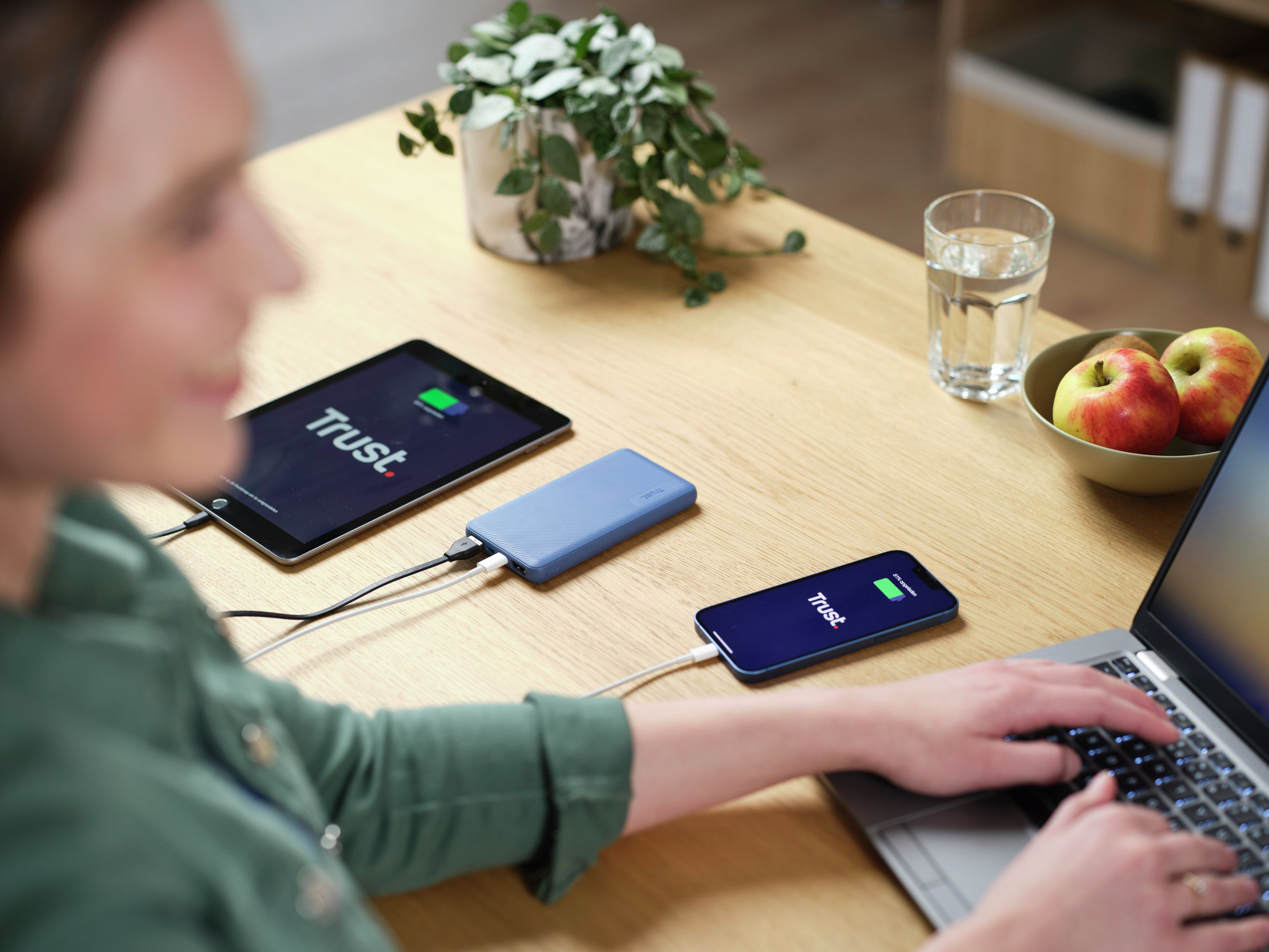 Eine Frau lädt Geräte auf: ein Tablet, ein Telefon und einen Laptop, alles auf einem Holztisch.