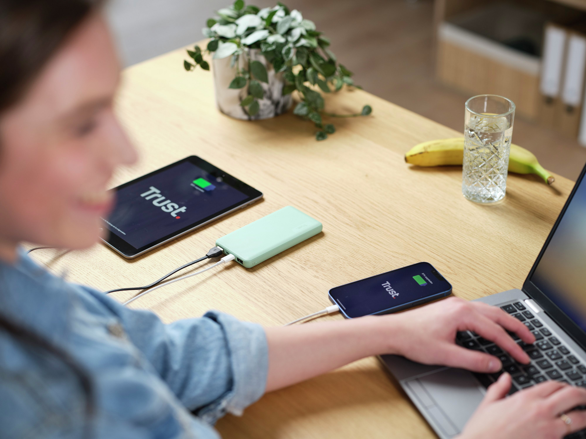 Een vrouw die een laptop gebruikt en apparaten oplaadt: een tablet en een telefoon, op een houten tafel.