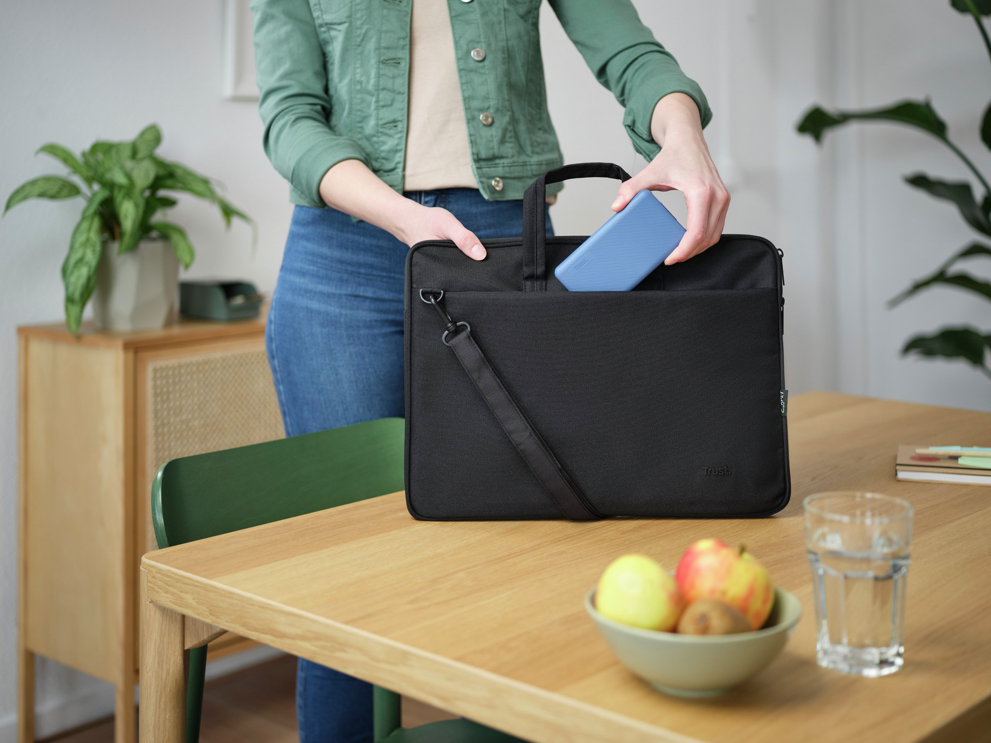 Eine Person legt einen blauen Laptop in eine schwarze Aktentasche. Andere Objekte liegen auf einem Holztisch.