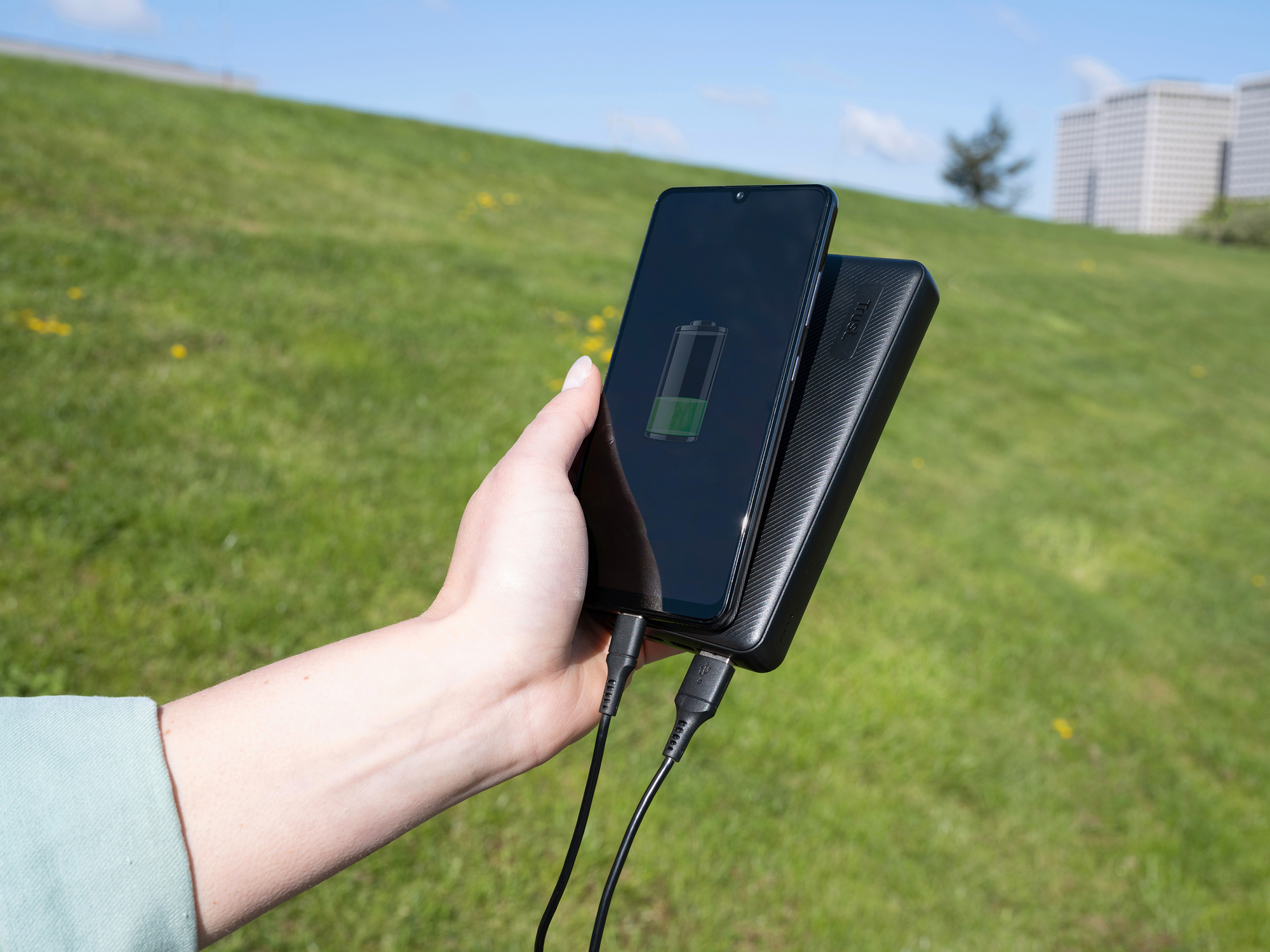 Ręka trzymająca telefon ładujący się z power banku na zewnątrz, tło trawy.