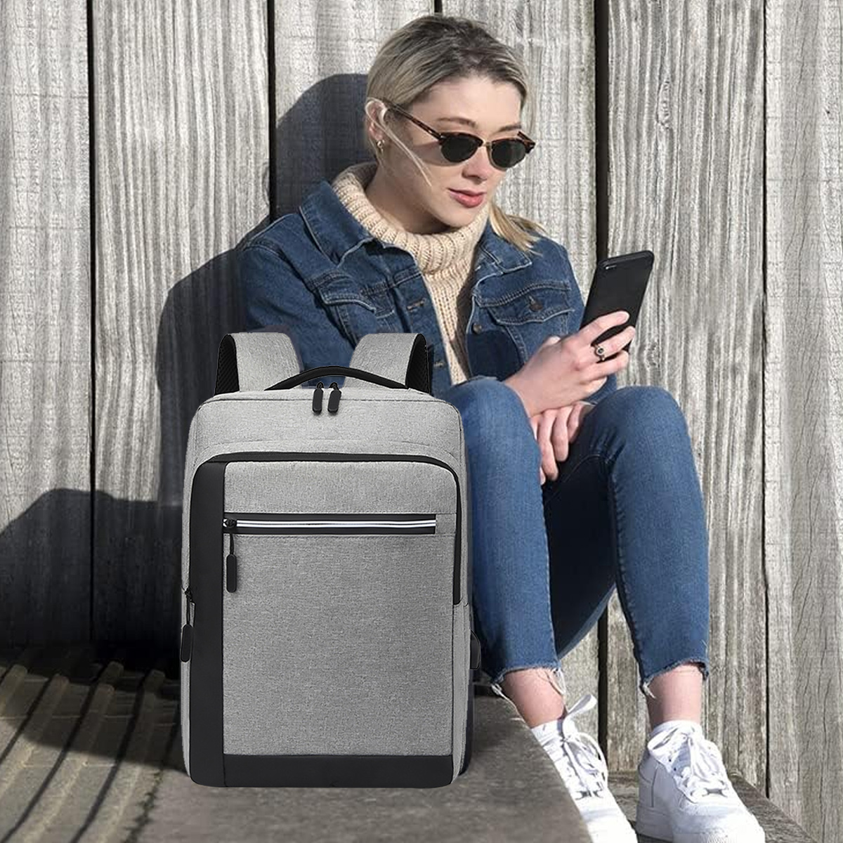 Eine Frau sitzt mit einem Rucksack. Sie trägt eine Sonnenbrille und schaut auf ihr Handy. Der Rucksack ist grau.