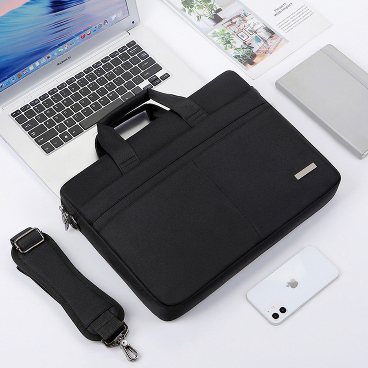 Schwarze Laptoptasche mit Laptop, Handy und Zubehör auf weißer Oberfläche. Die Tasche hat einen Schultergurt.