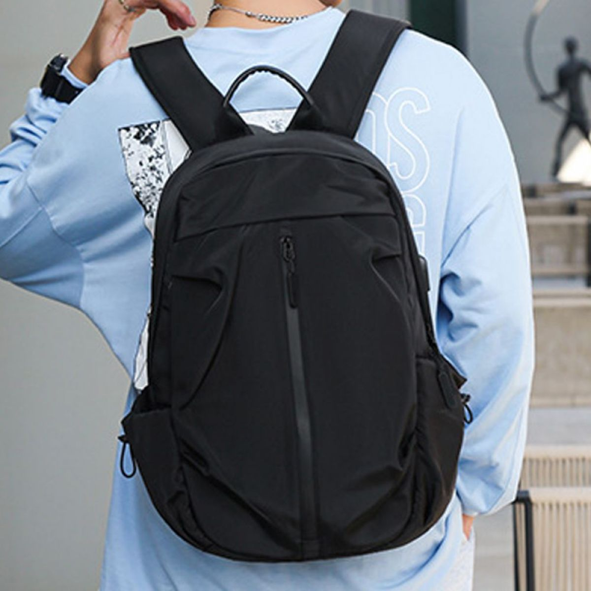 Schwarzer Rucksack mit Fronttasche, getragen von einer Person in blauem Hemd, im Freien.