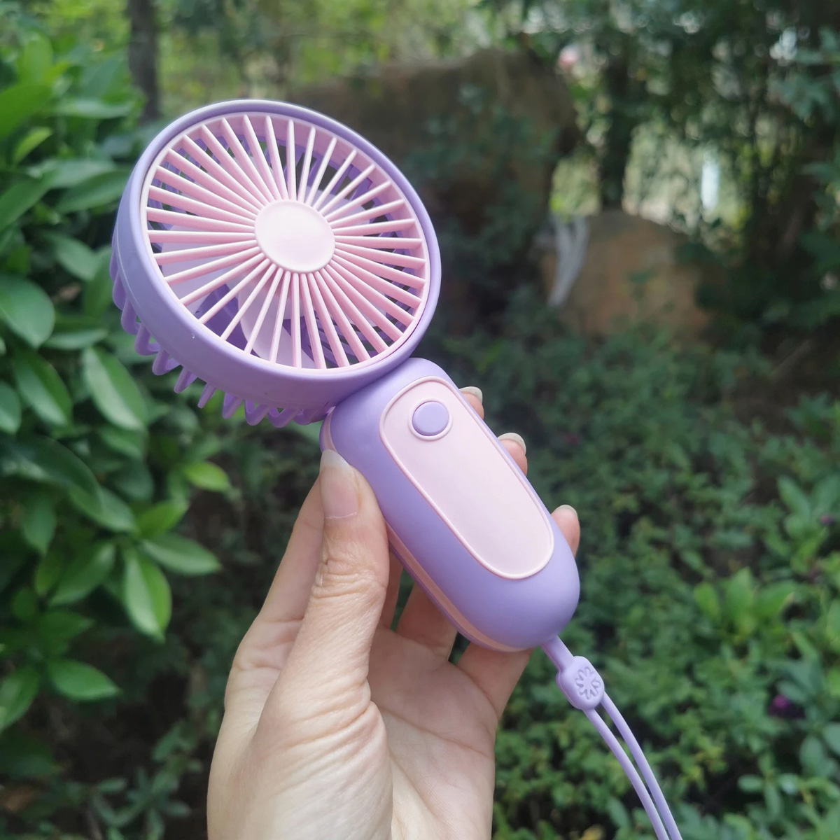 Mano sosteniendo un mini ventilador lila y rosa. Follaje verde al fondo.
