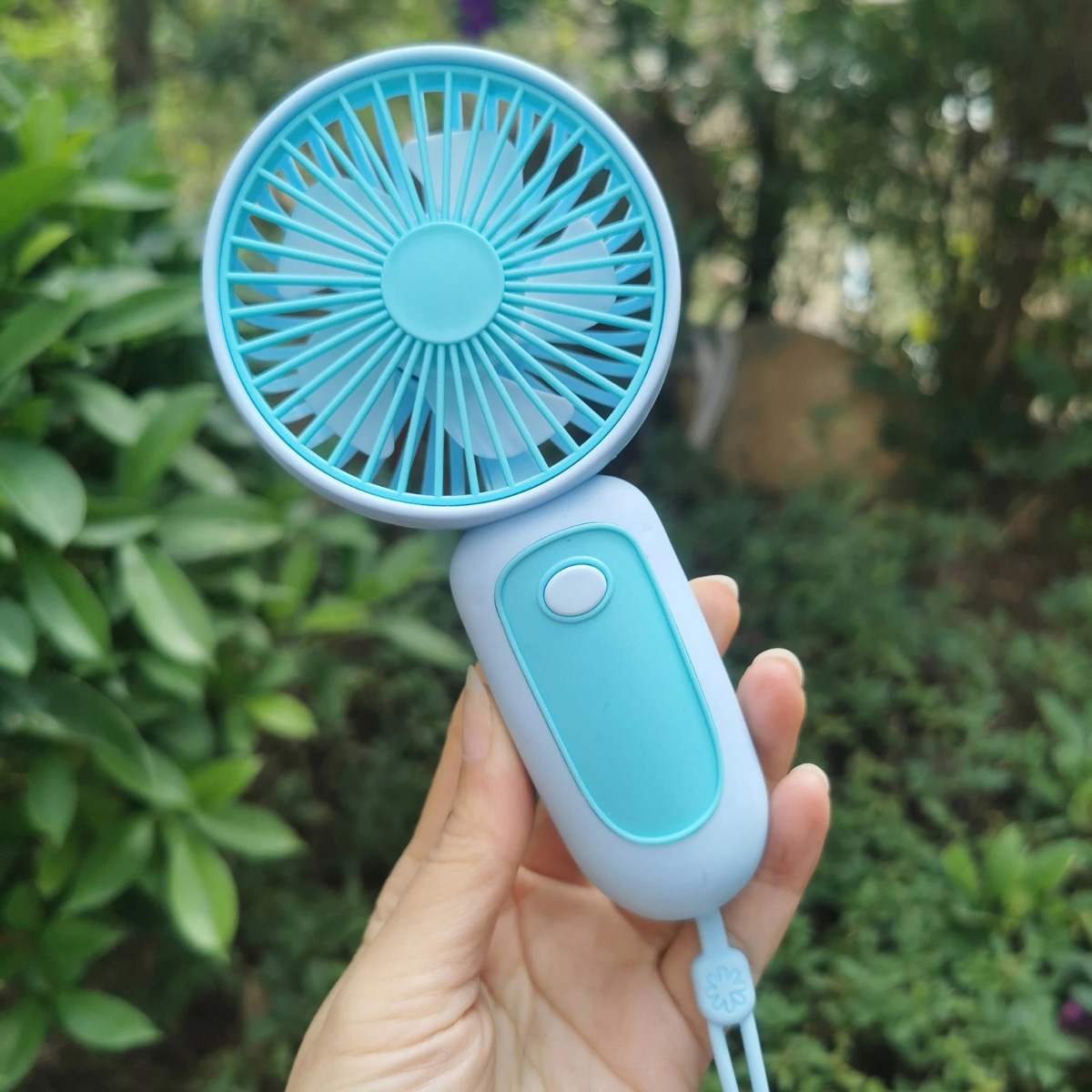 Una mano sostiene un ventilador portátil azul claro con aspas color turquesa, sobre un fondo verde y frondoso.