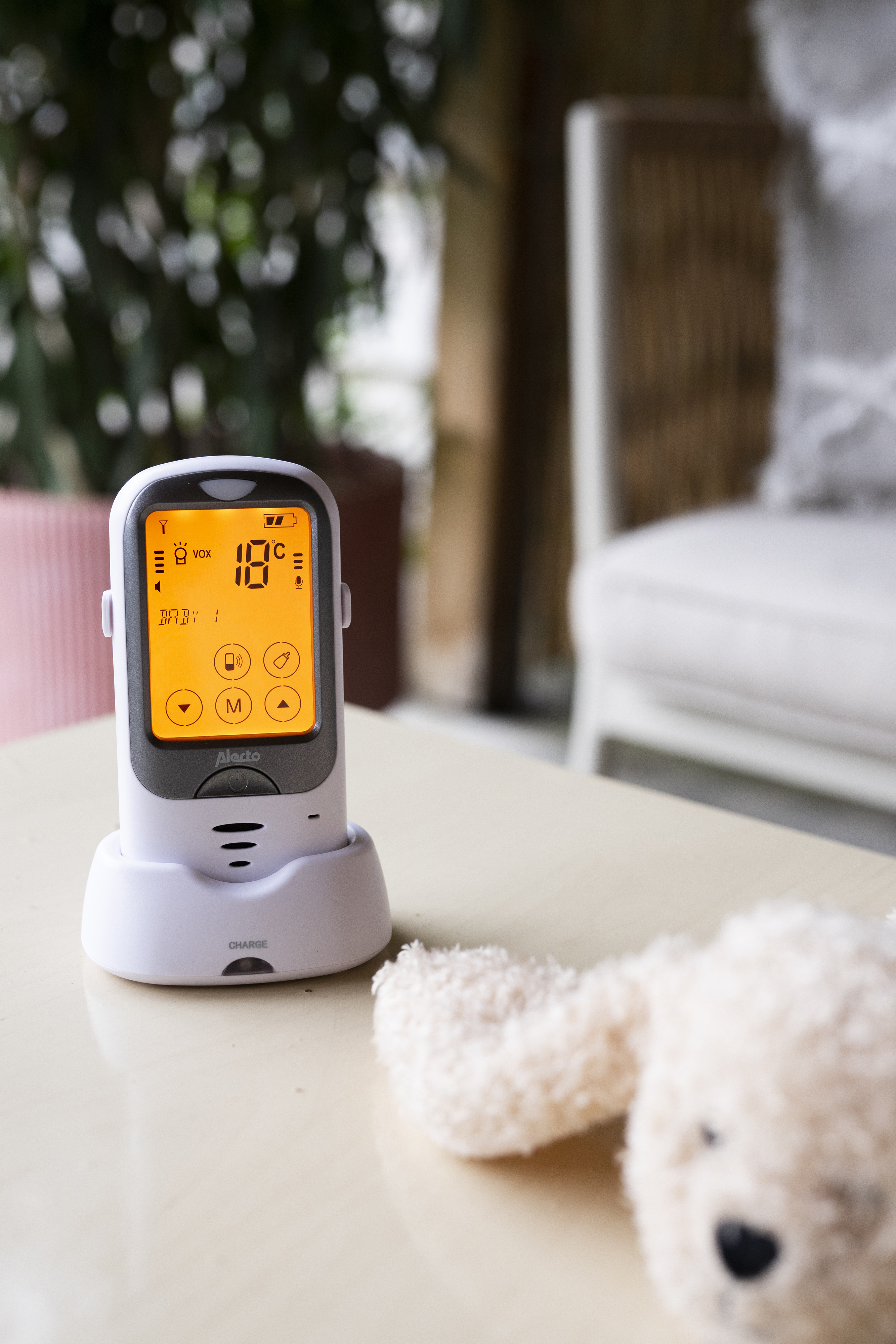 A white baby monitor on a table. It shows the temperature is 18 degrees. A teddy bear is next to it.