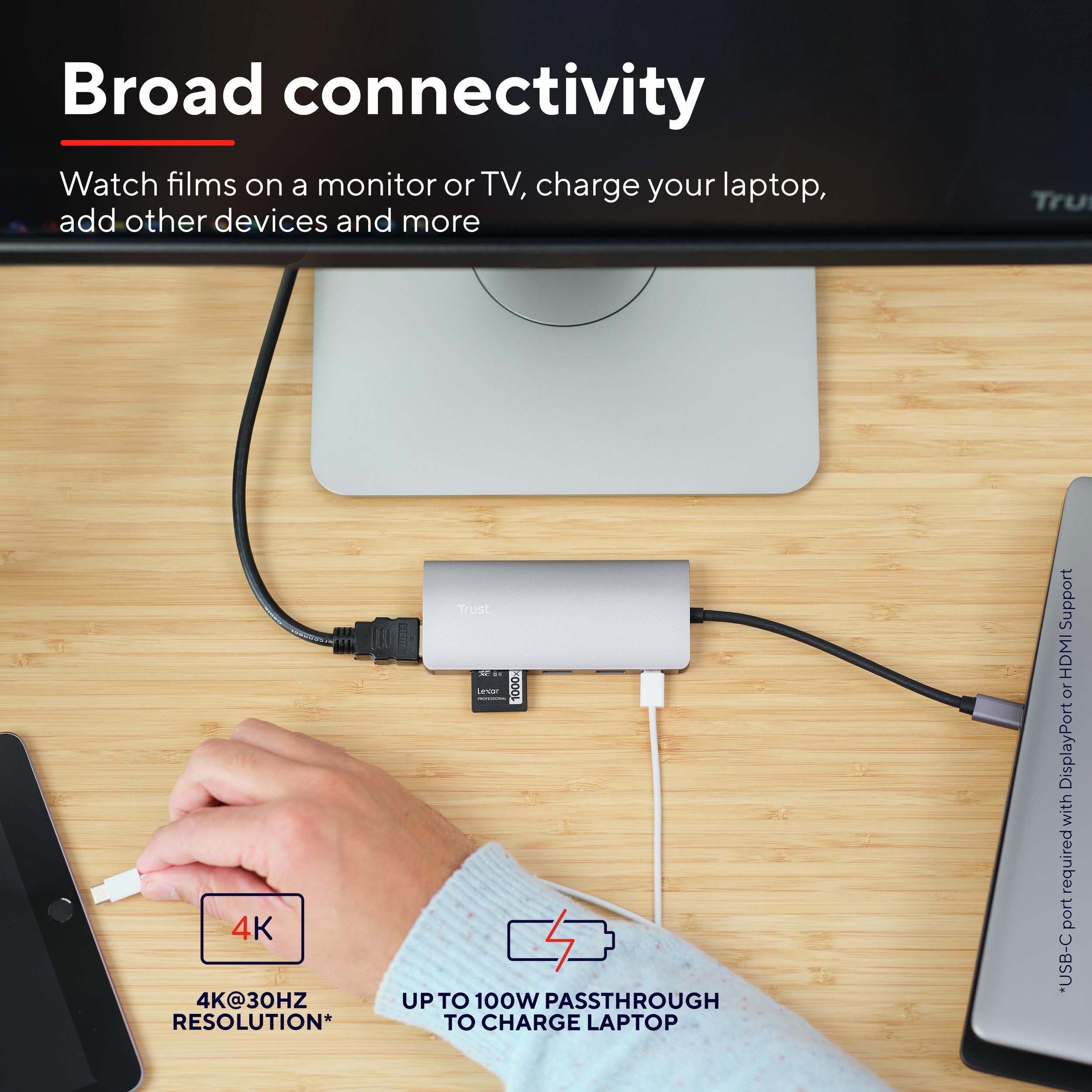 Eine Hand steckt ein USB-C-Kabel in einen silbernen Hub auf einer Holzoberfläche. Ein iPad, Laptop sind sichtbar.