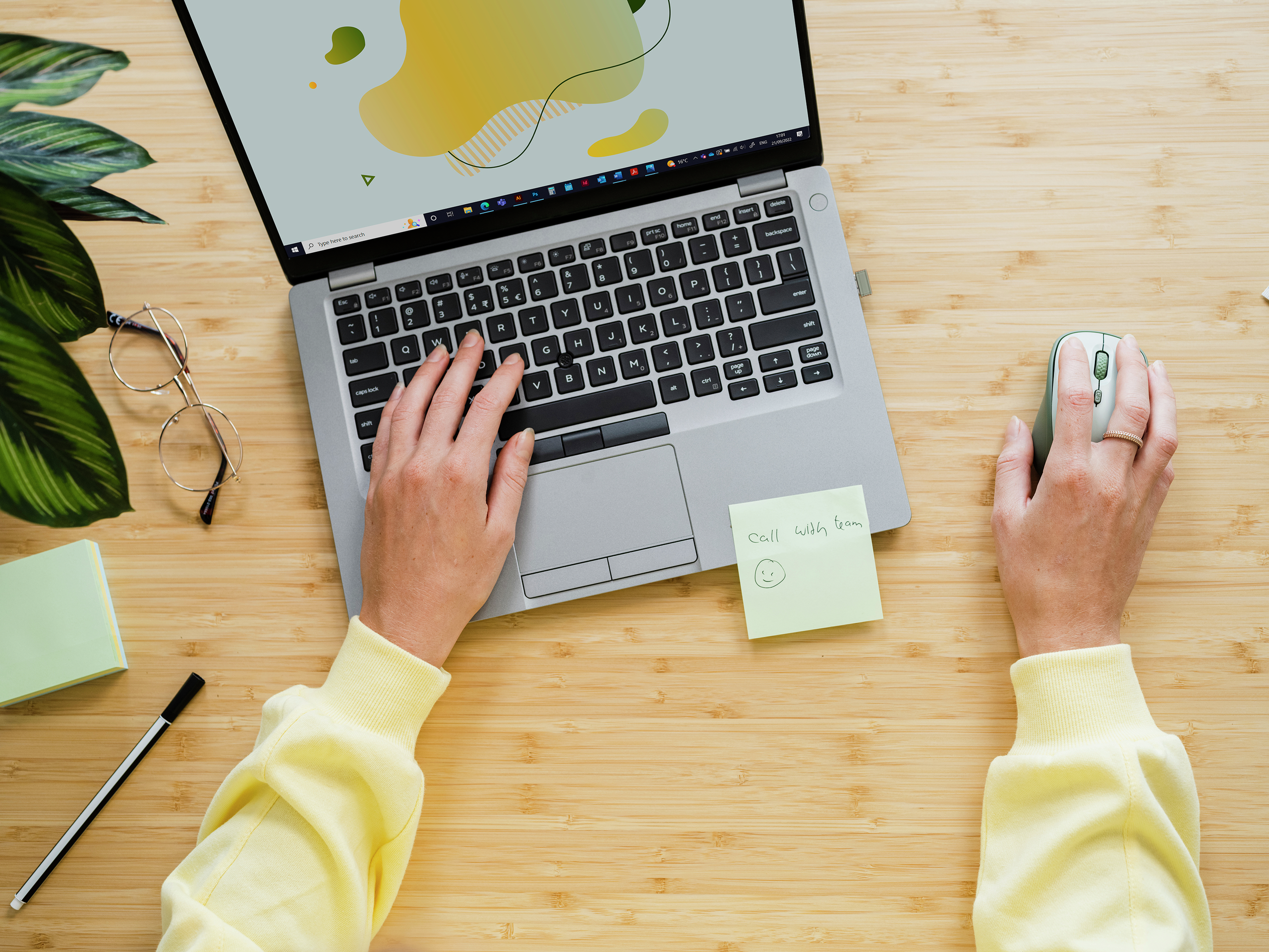 Widok z góry na osobę piszącą na laptopie, z karteczką samoprzylepną i długopisem na drewnianym biurku.