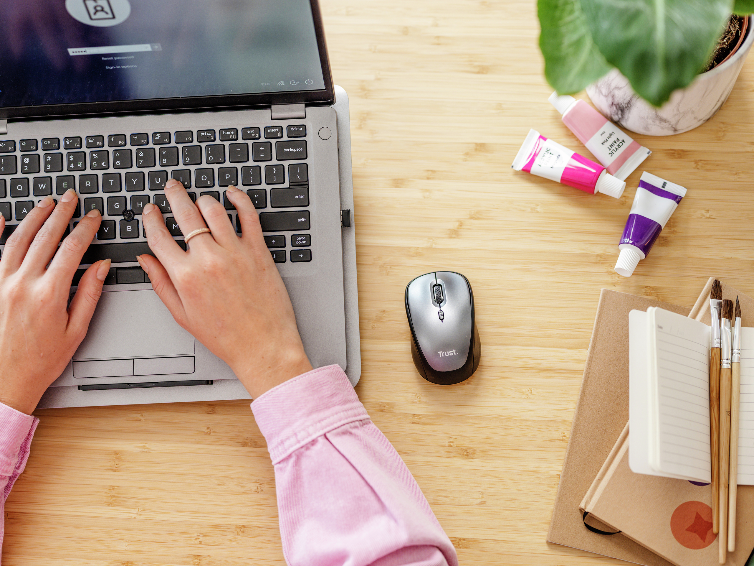 Ręce piszące na klawiaturze laptopa z myszą i materiałami artystycznymi.