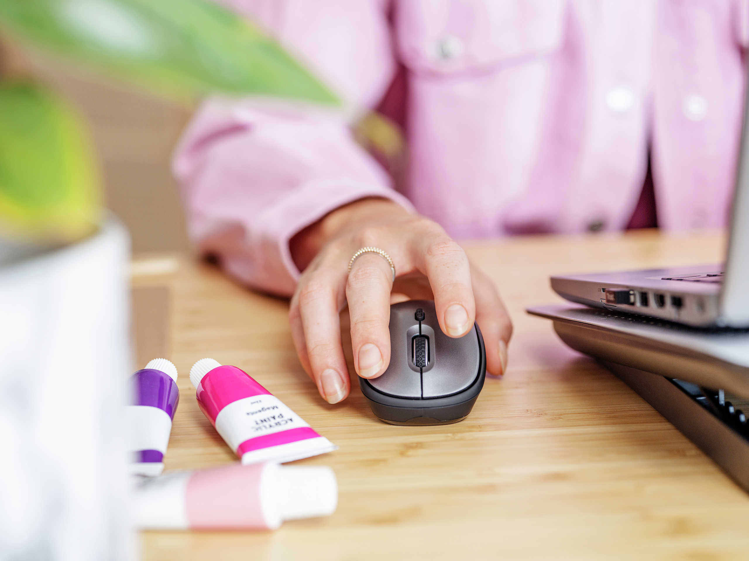 Dłoń używająca szarej myszy na drewnianym biurku. Różowa kurtka i tubki z farbą, widoczny laptop.