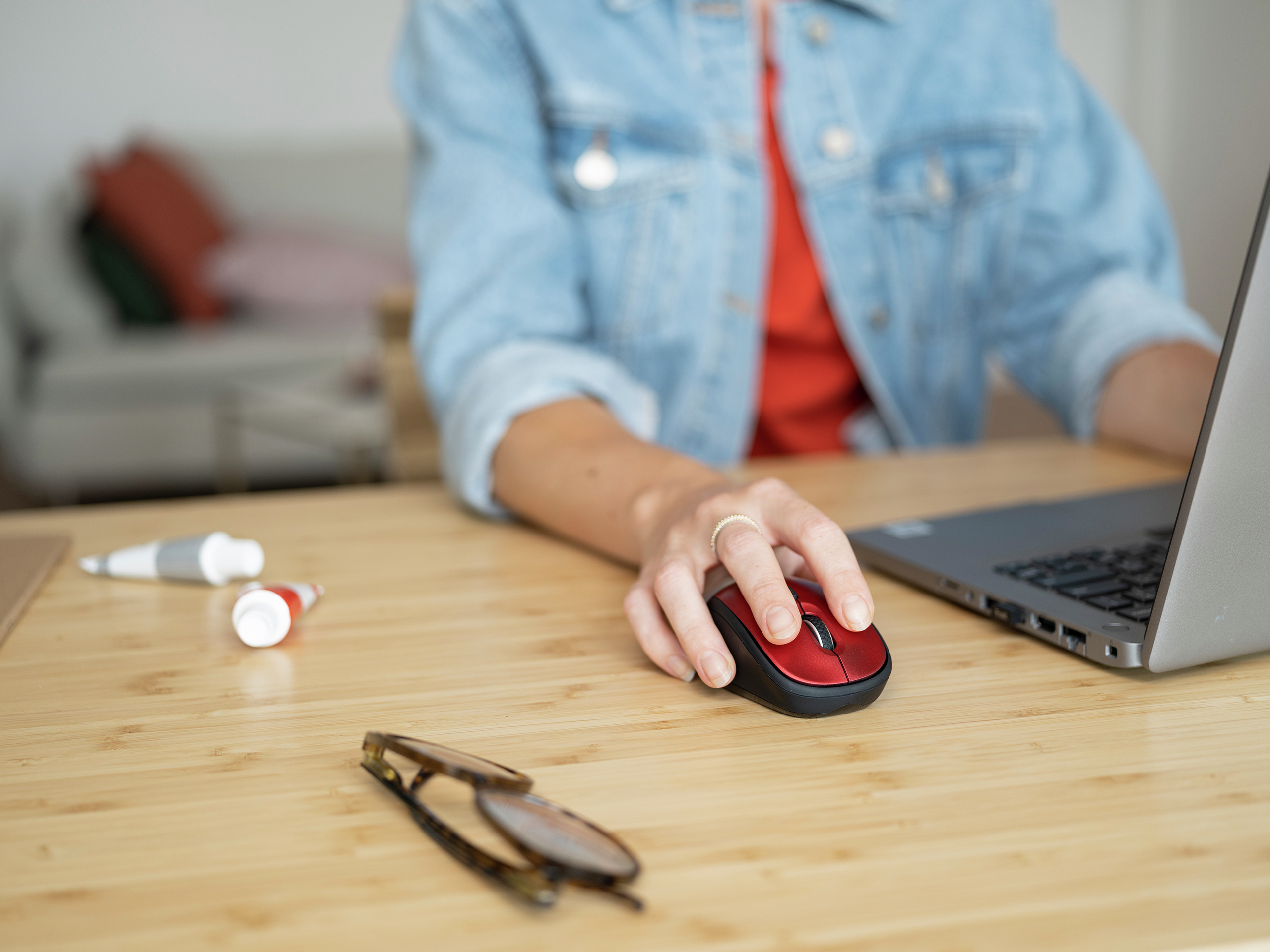 Ręka osoby używającej czerwonej myszy na drewnianym biurku. Okulary i tubki z farbą na biurku.