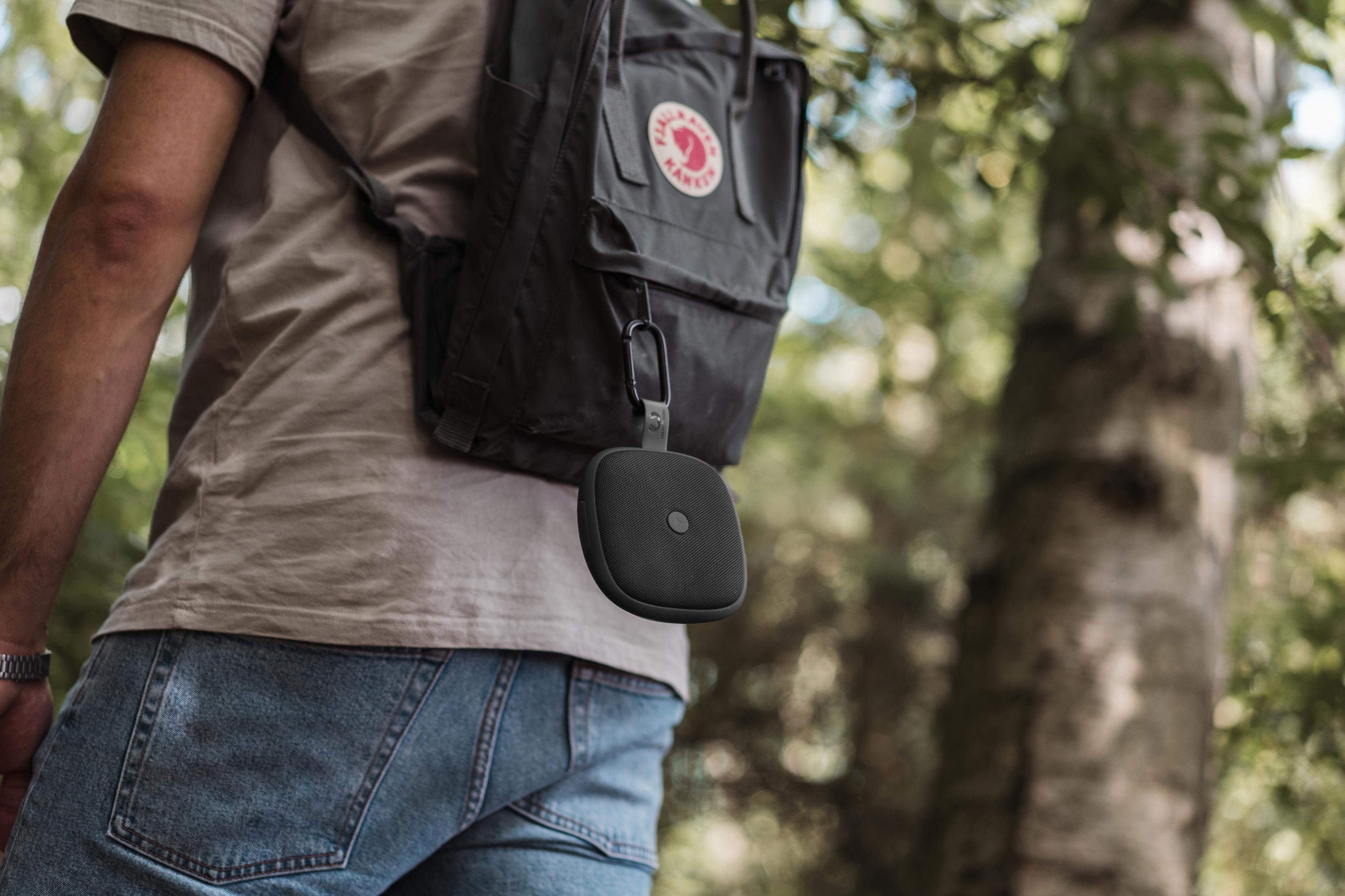 Osoba niosąca plecak z przymocowanym do niego czarnym głośnikiem podczas spaceru na łonie natury.