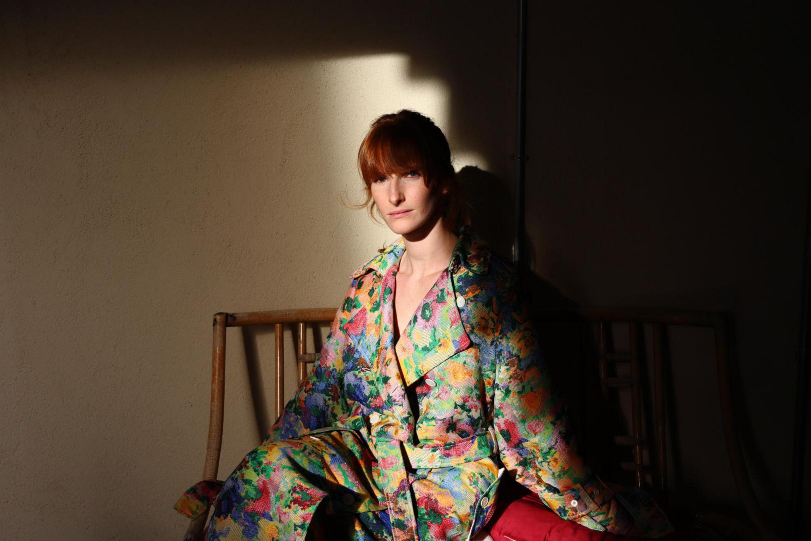 Woman in colorful patterned jacket sitting on a wooden bench. Sunlight on the scene.