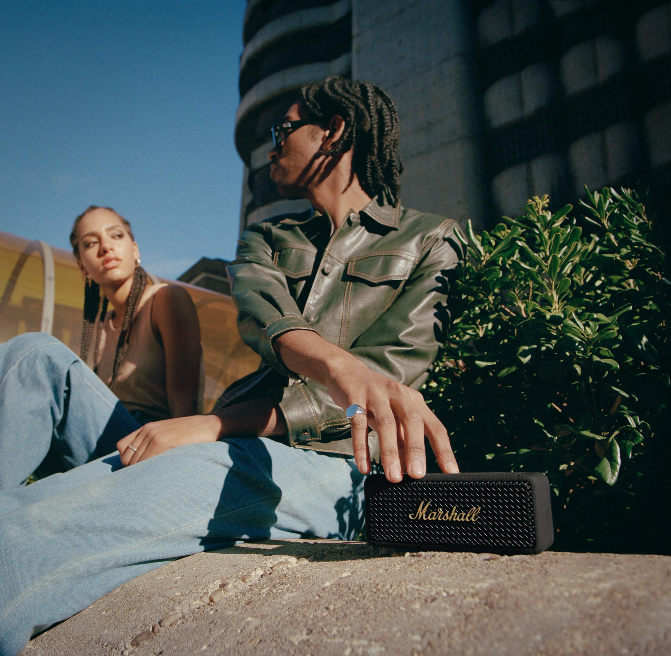 Dos personas sentadas al aire libre con un altavoz negro Marshall. Una persona sostiene el altavoz.