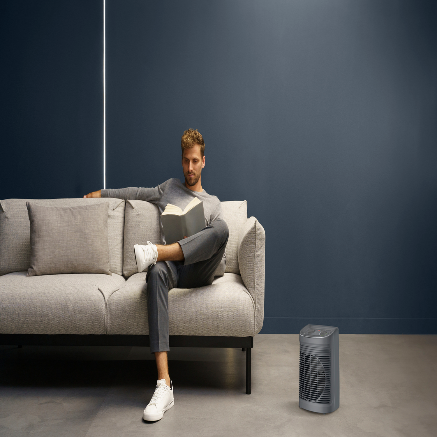 Mężczyzna czyta książkę na kanapie, obok szarego grzejnika. Ciemnoniebieska ściana w tle.