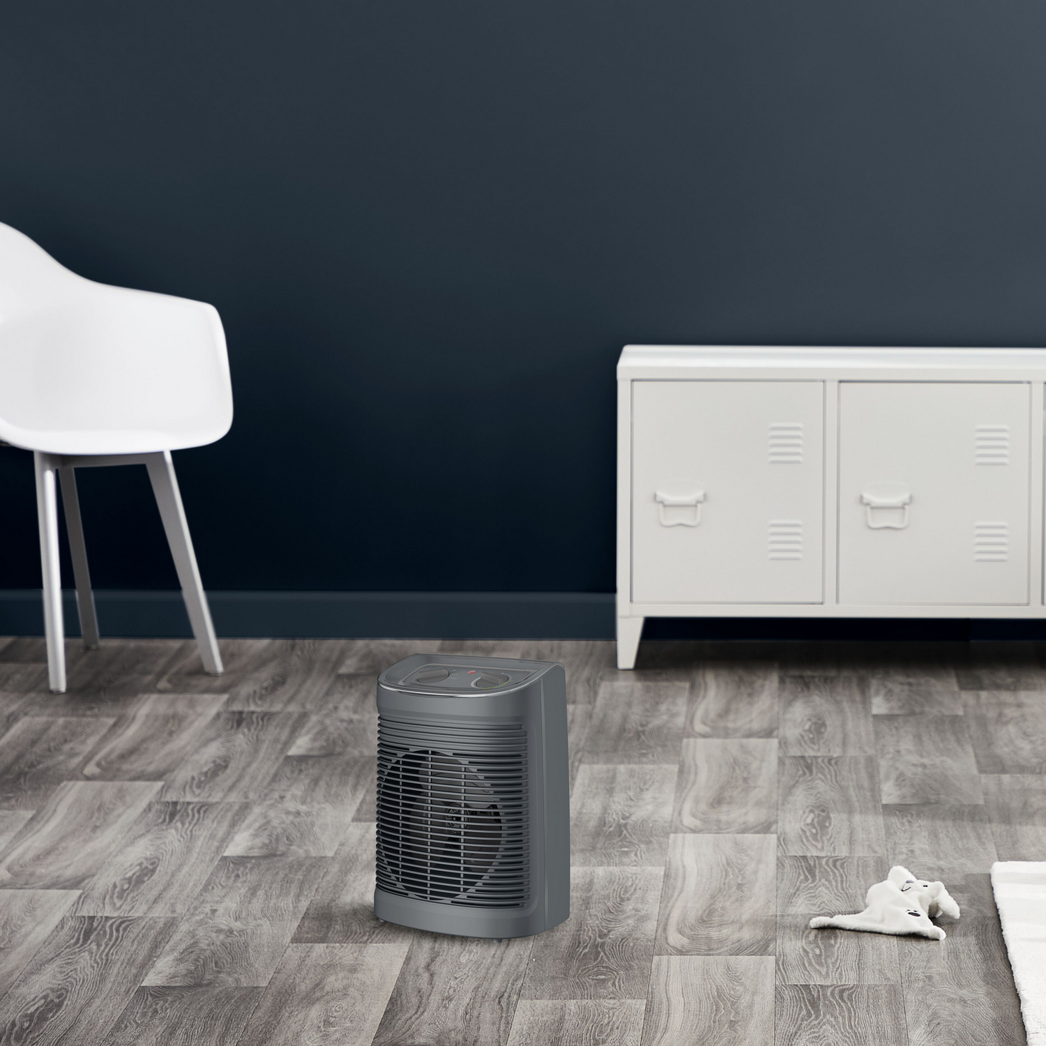 Szary grzejnik elektryczny na drewnianej podłodze. Białe krzesło i szafka w tle.