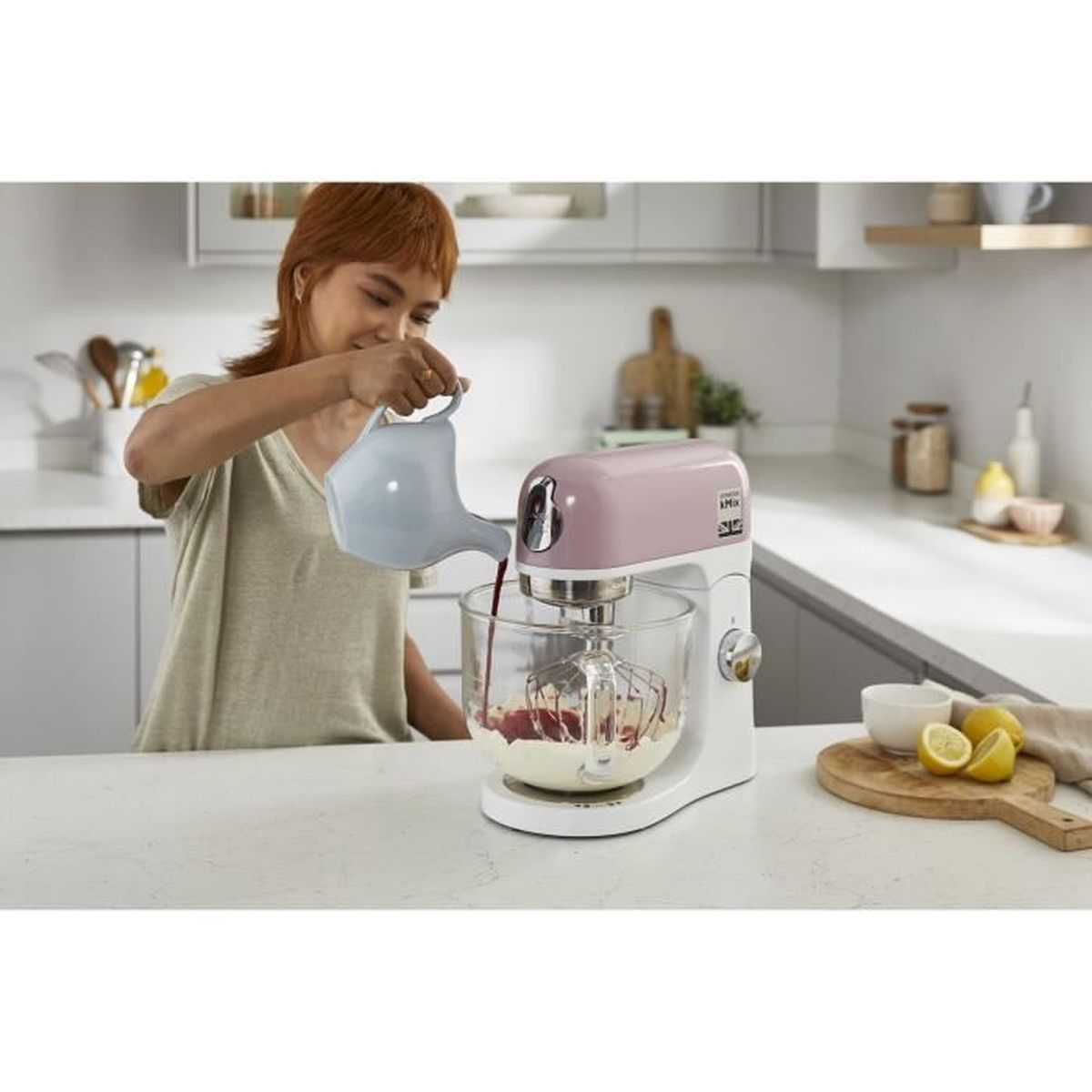 Donna che versa liquido in un'impastatrice rosa e bianca. Limoni su tagliere in legno. Ambiente cucina.
