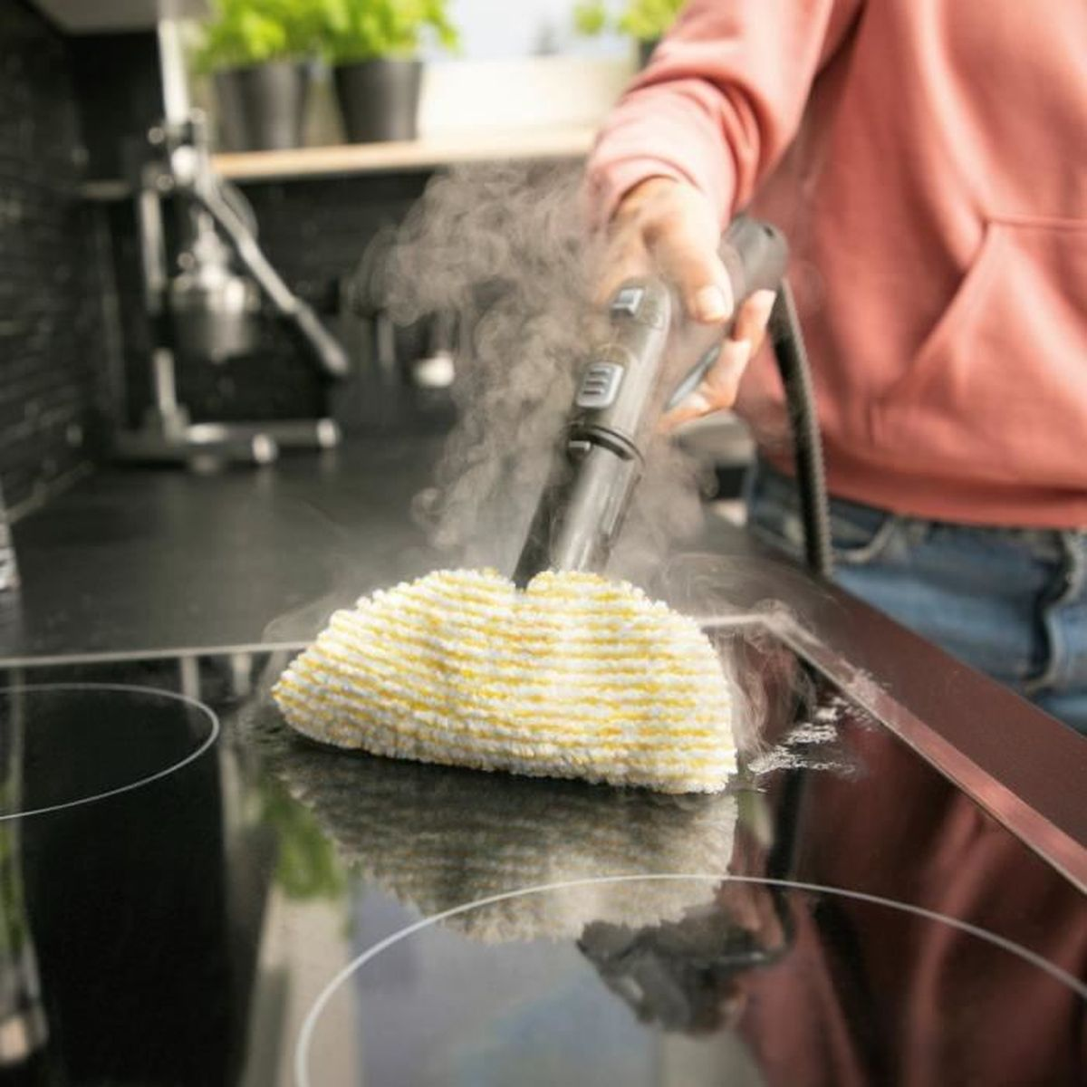 Una persona usa un pulitore a vapore con panno giallo su piano cottura in vetroceramica nero.