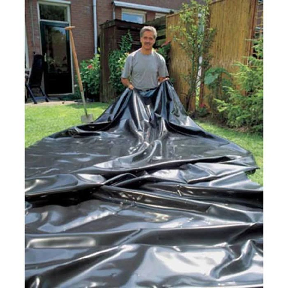 Ein Mann hält eine große, schwarze Plastikfolie in einem Garten und bereitet die Installation eines Teichs vor.
