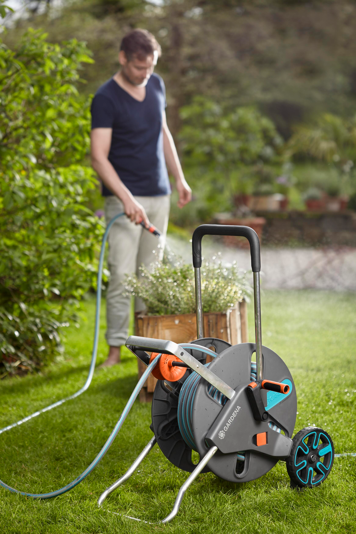Mann benutzt eine Gartenschlauchhaspel. Grünes Gras, hölzerner Pflanzkasten im Hintergrund.