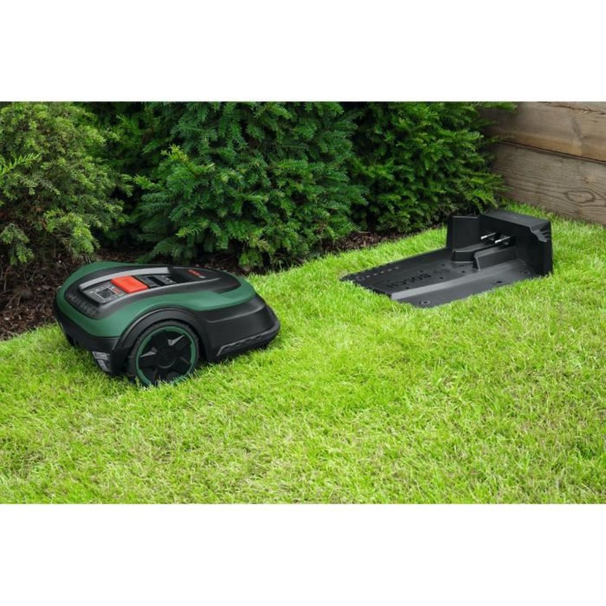 Grüner und schwarzer Roboter-Rasenmäher auf Gras mit Ladestation, grüne Büsche im Hintergrund.