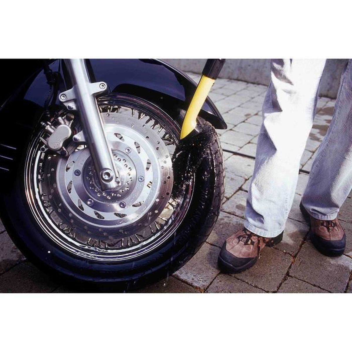 Osoba czyszcząca oponę motocyklową żółtą szczotką, na ceglanej powierzchni.