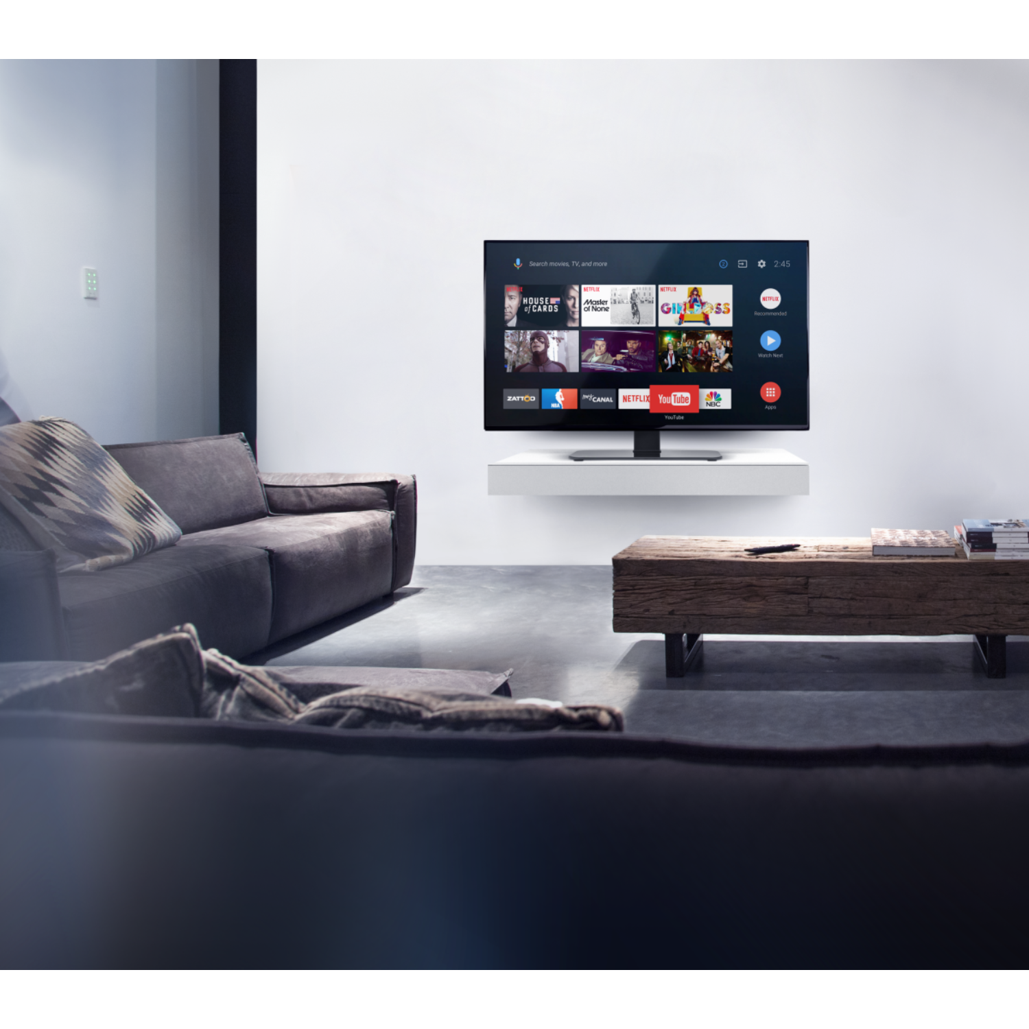 Woonkamer met tv op witte plank, houten tafel en grijze bank.
