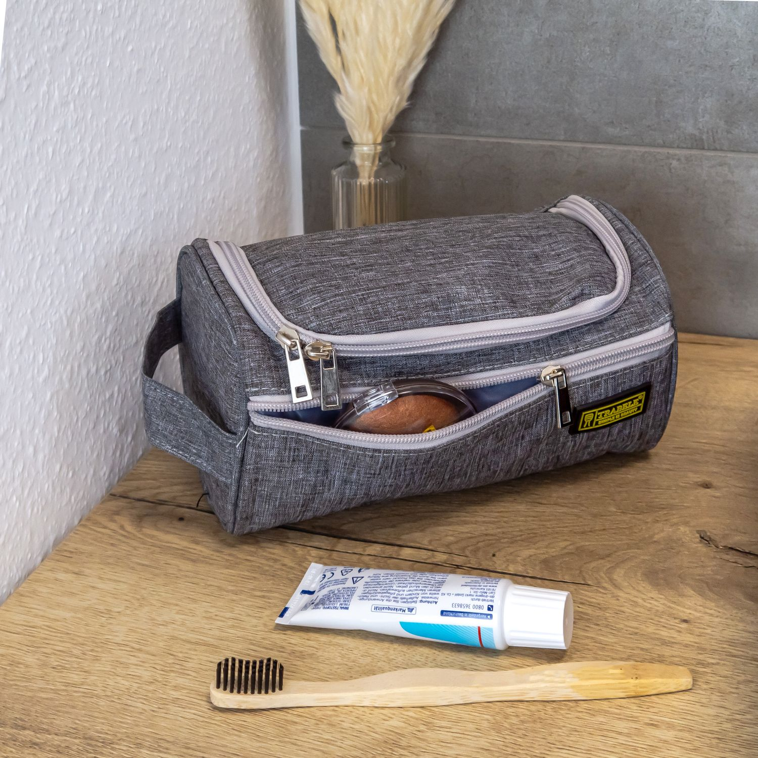 Graue Kosmetiktasche mit offenem Reißverschluss. Zahnbürste und Zahnpasta liegen auf der Theke.