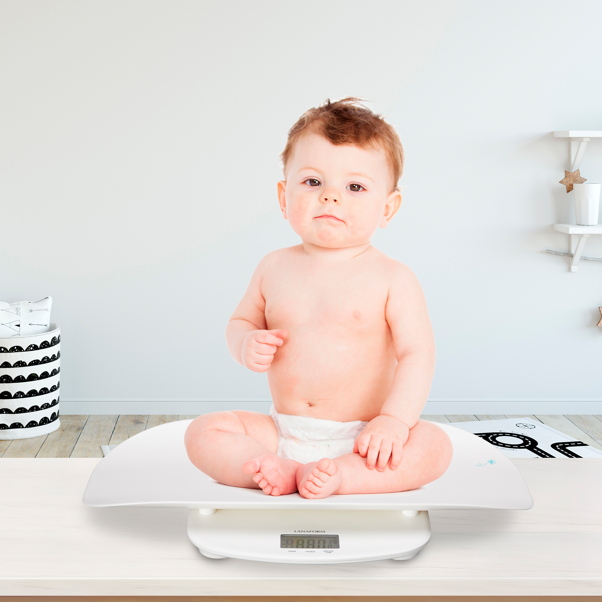 Een baby zit op een witte weegschaal. Achtergrond: muur en decoratieve items.