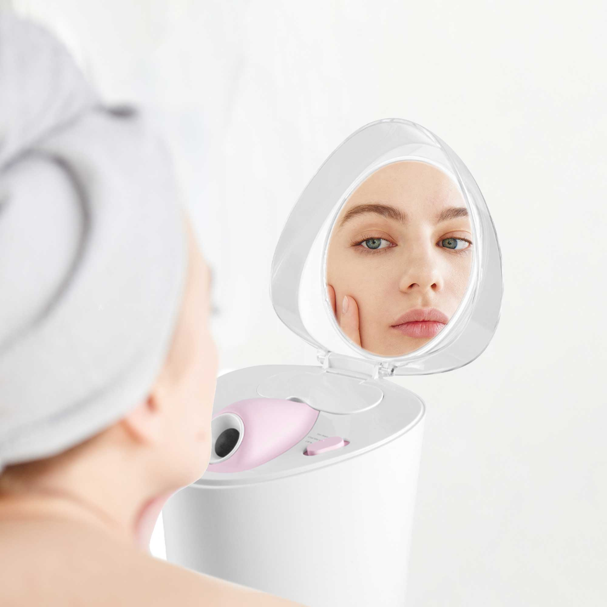 Woman looking in a mirror above a facial steamer. The woman is wearing a towel on her head.