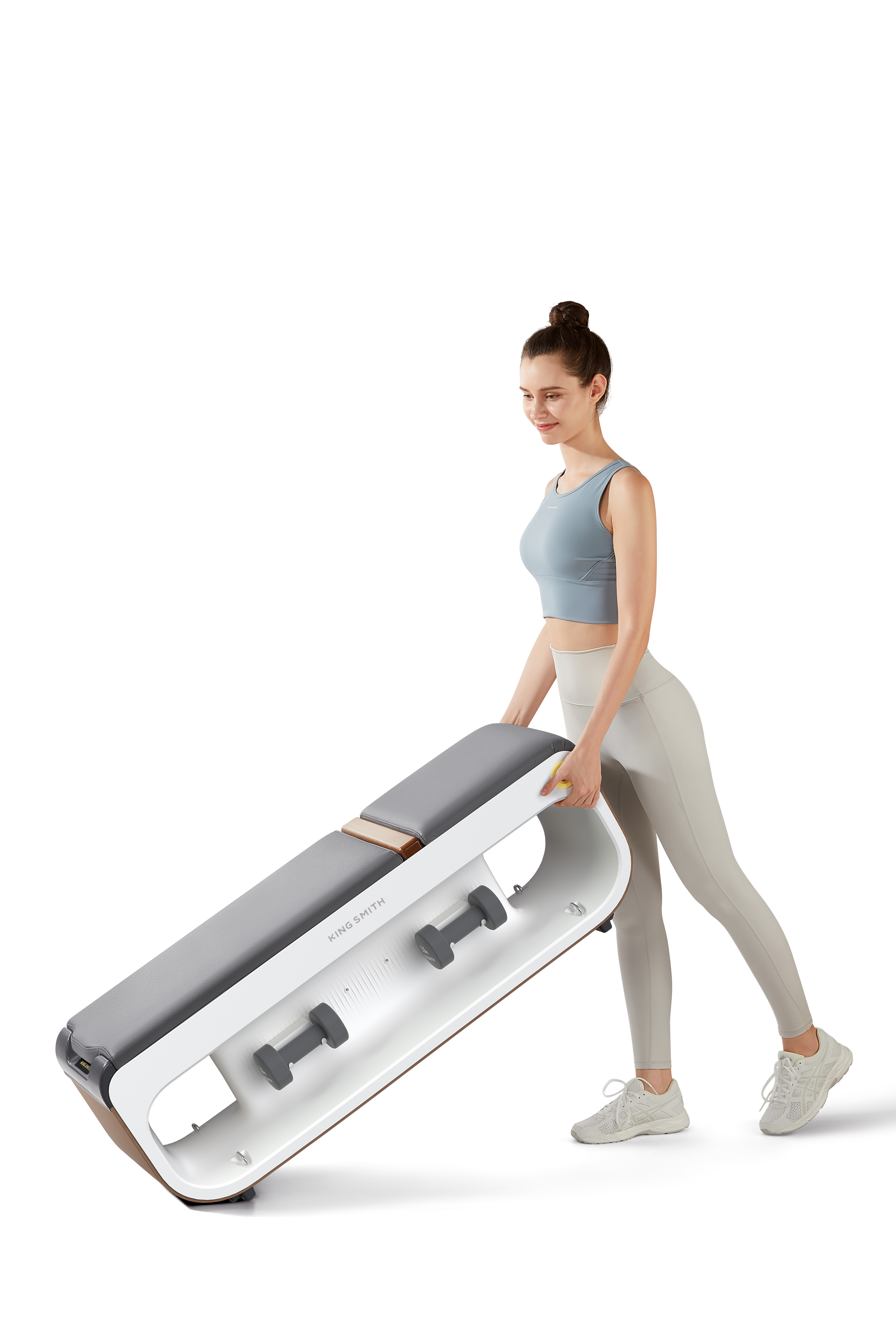 Kobieta niesie biało-szarą ławkę do ćwiczeń z dołączonymi hantlami. Uśmiecha się i ma na sobie strój sportowy.