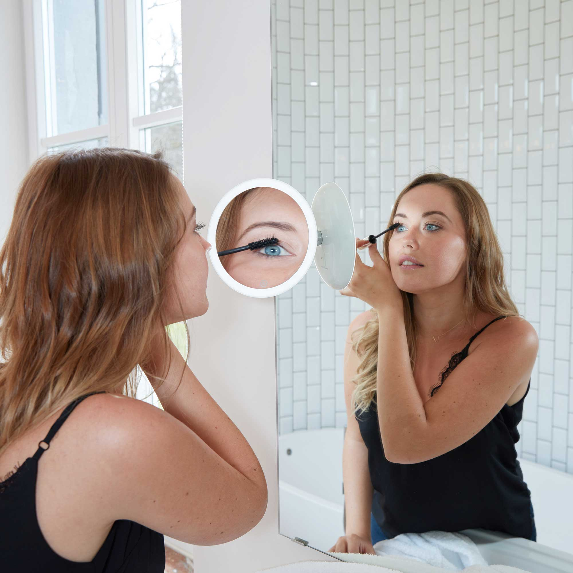 Frau, die Mascara in einem Spiegel aufträgt, mit einem Vergrößerungsspiegel.
