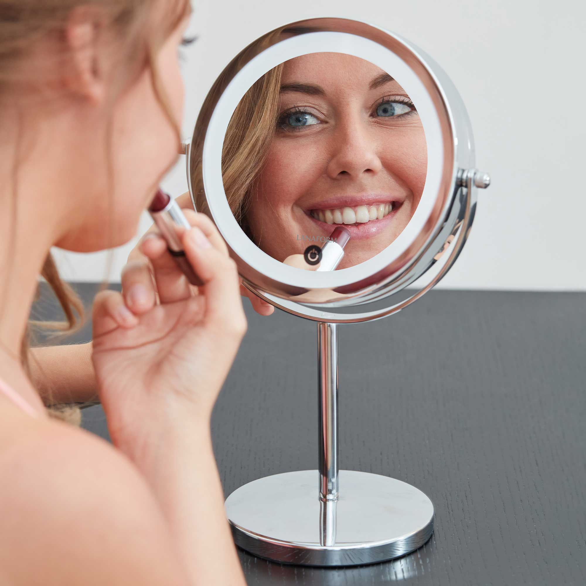 Woman applying lipstick in a lighted, round vanity mirror. The mirror reflects her face. Dark table.