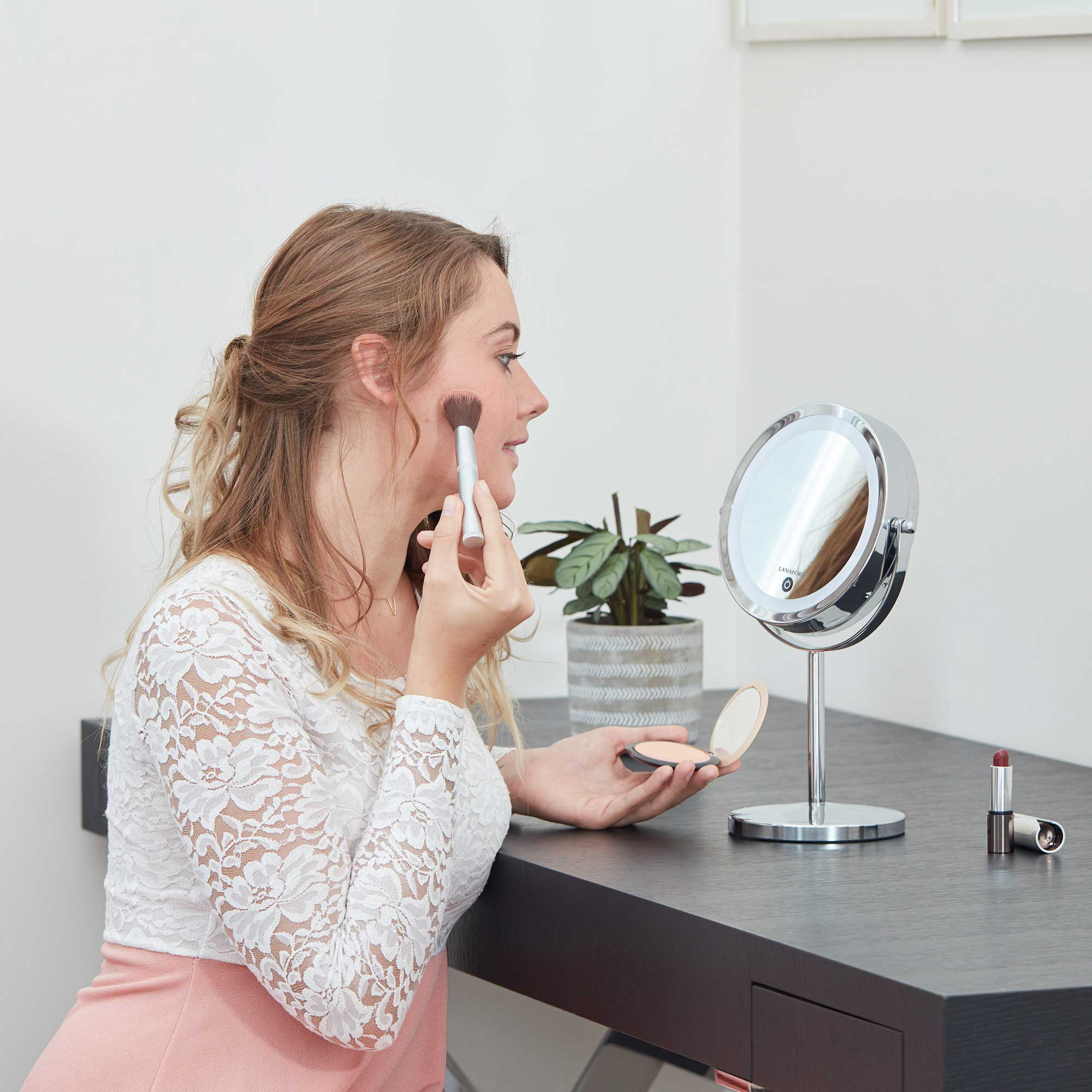 A woman applying makeup with a brush, in front of a mirror. The background is a dark table.