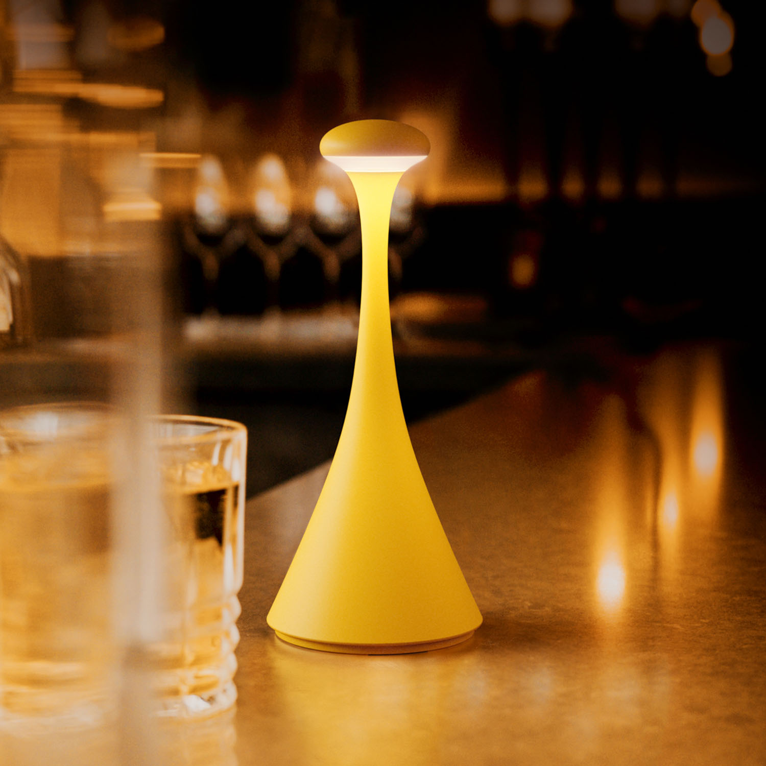 Gelbe, pilzförmige Lampe auf einem Tisch. Ein Glas und verschwommener Hintergrund sind sichtbar.