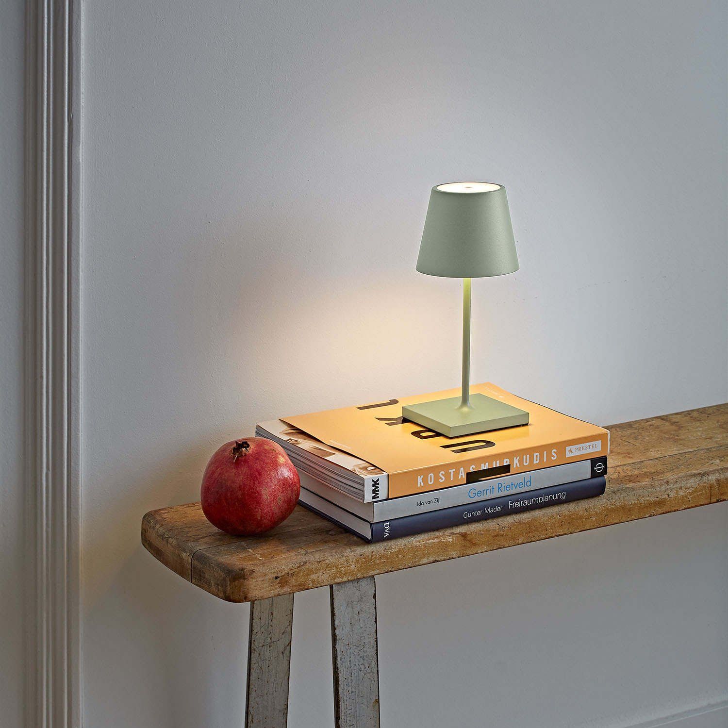 Eine grüne Tischlampe auf Büchern mit einem Granatapfel auf einer Holzbank vor einer weißen Wand.