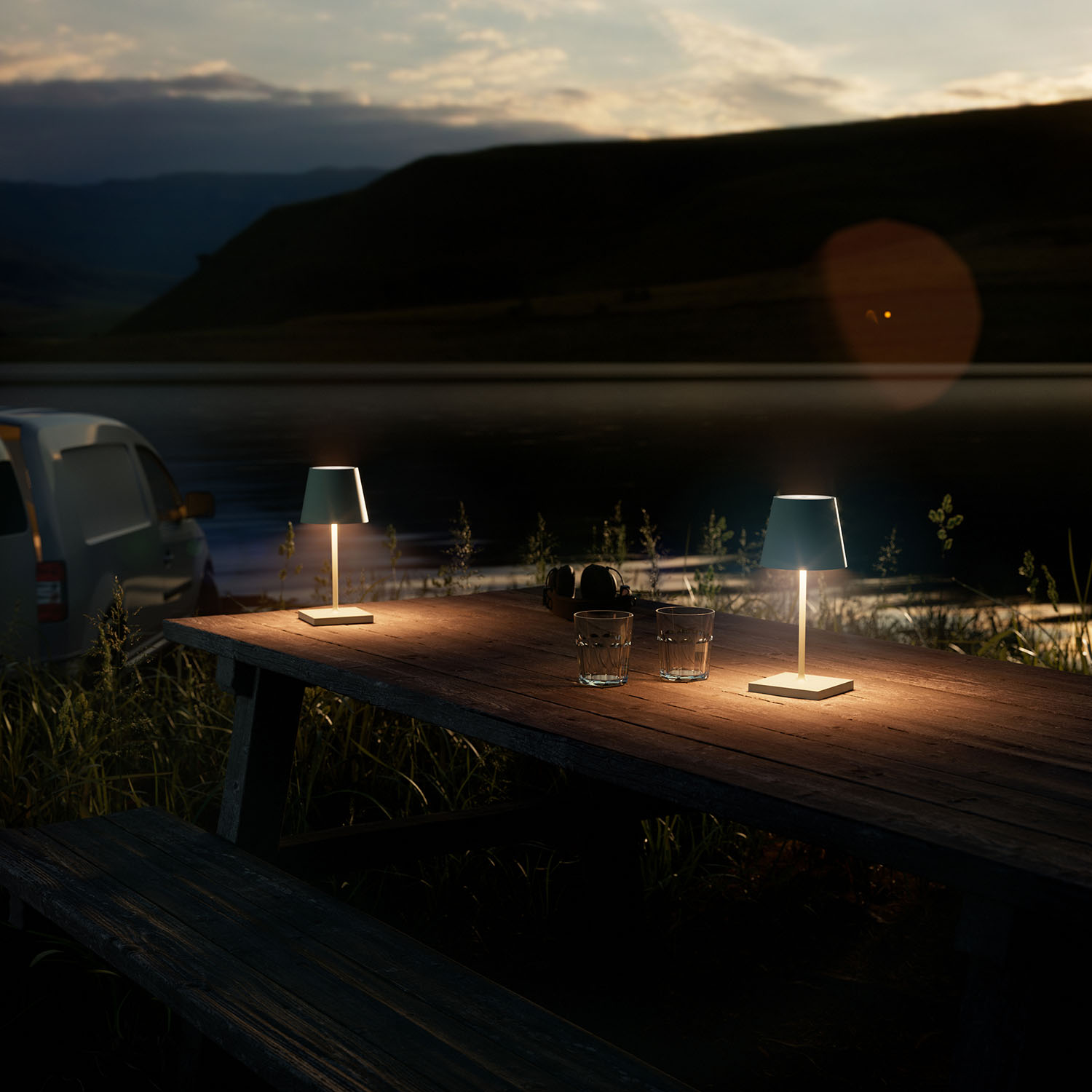 Zwei Lampen auf einem Holztisch in der Abenddämmerung am See, mit Auto im Hintergrund.