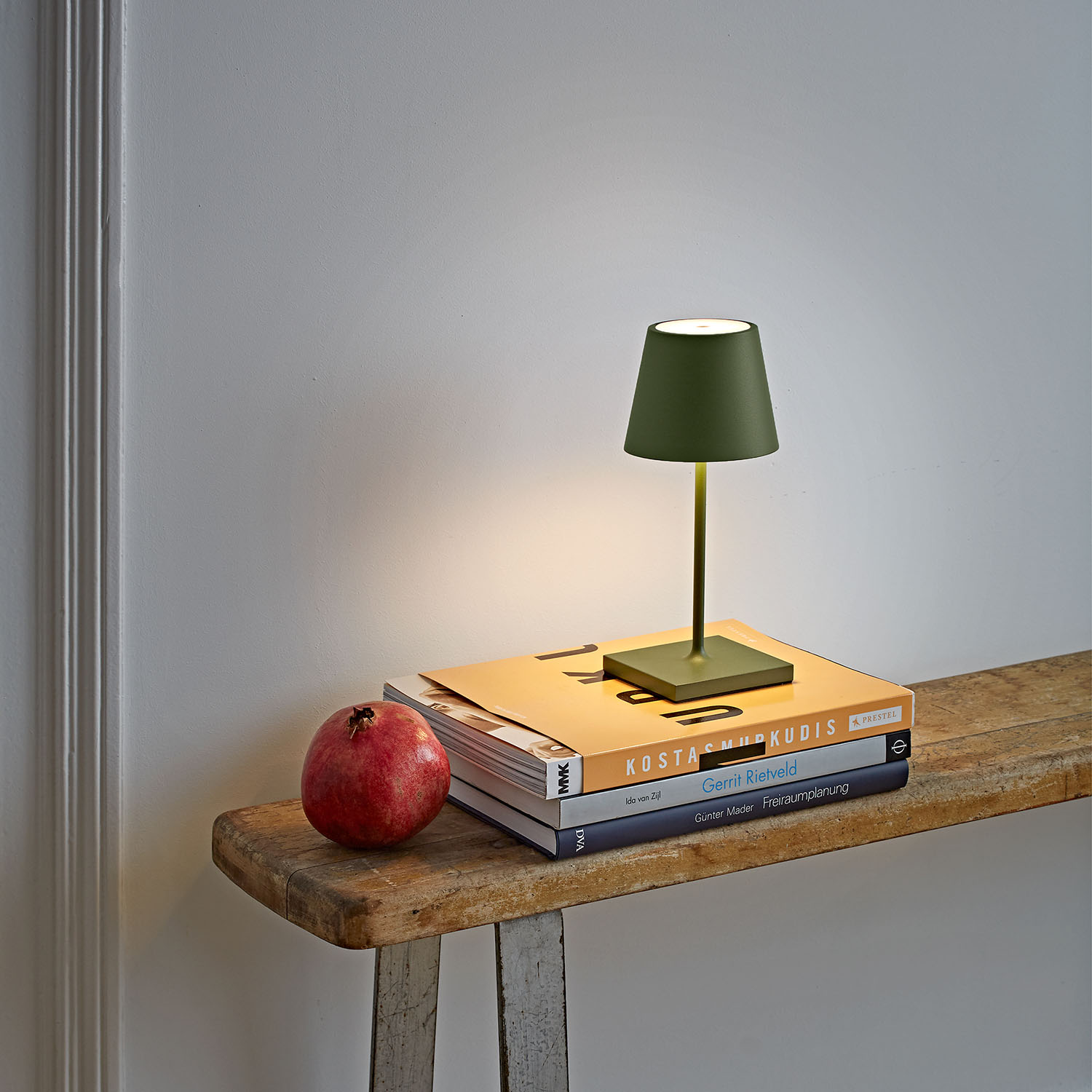 Eine grüne Lampe auf einem Stapel Bücher, mit einem Granatapfel auf einer Holzoberfläche. Weiße Wand im Hintergrund.