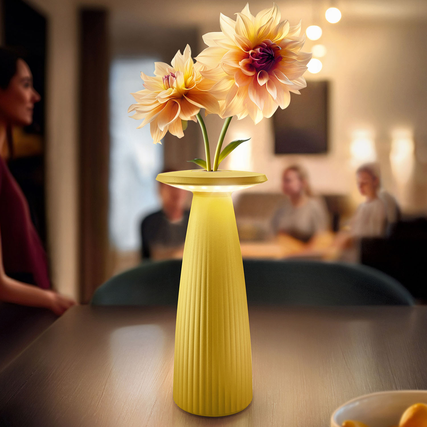 Gelbe Lampe mit zwei Blumen auf einem Tisch. Personen sind unscharf im Hintergrund.