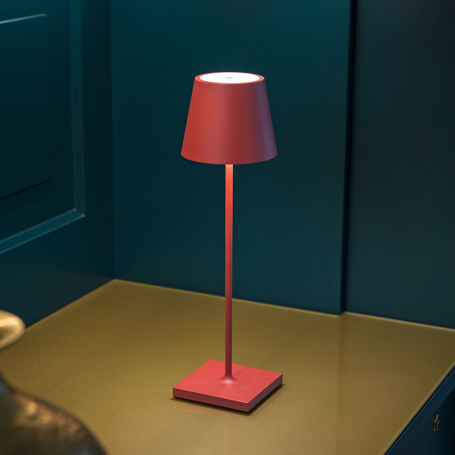 Rote Tischlampe mit quadratischem Sockel. Die Lampe befindet sich auf einer Oberfläche mit grünem Hintergrund.