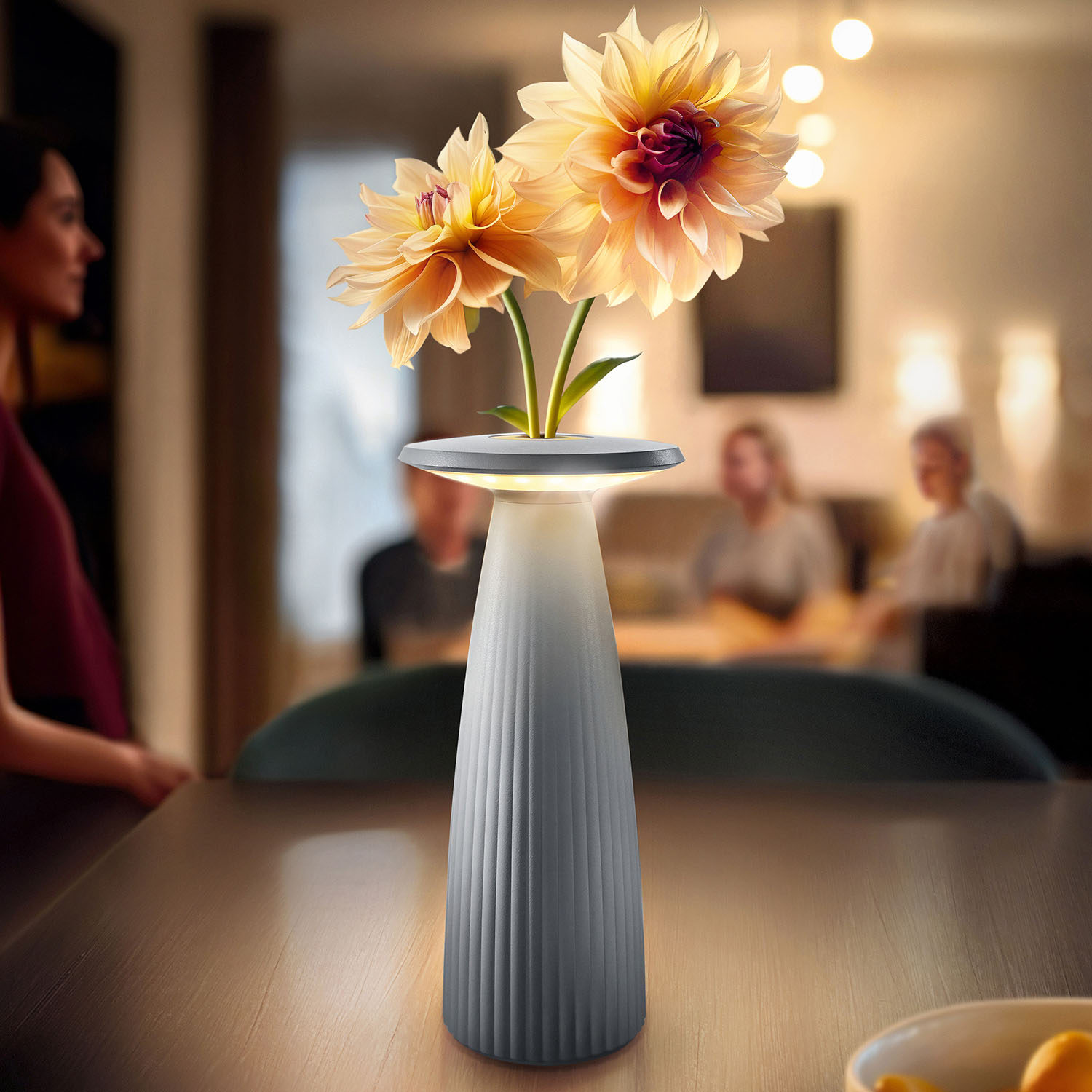Eine moderne Vase mit zwei Blumen auf einem Tisch. Personen sind im Hintergrund unscharf. Weiche Beleuchtung.