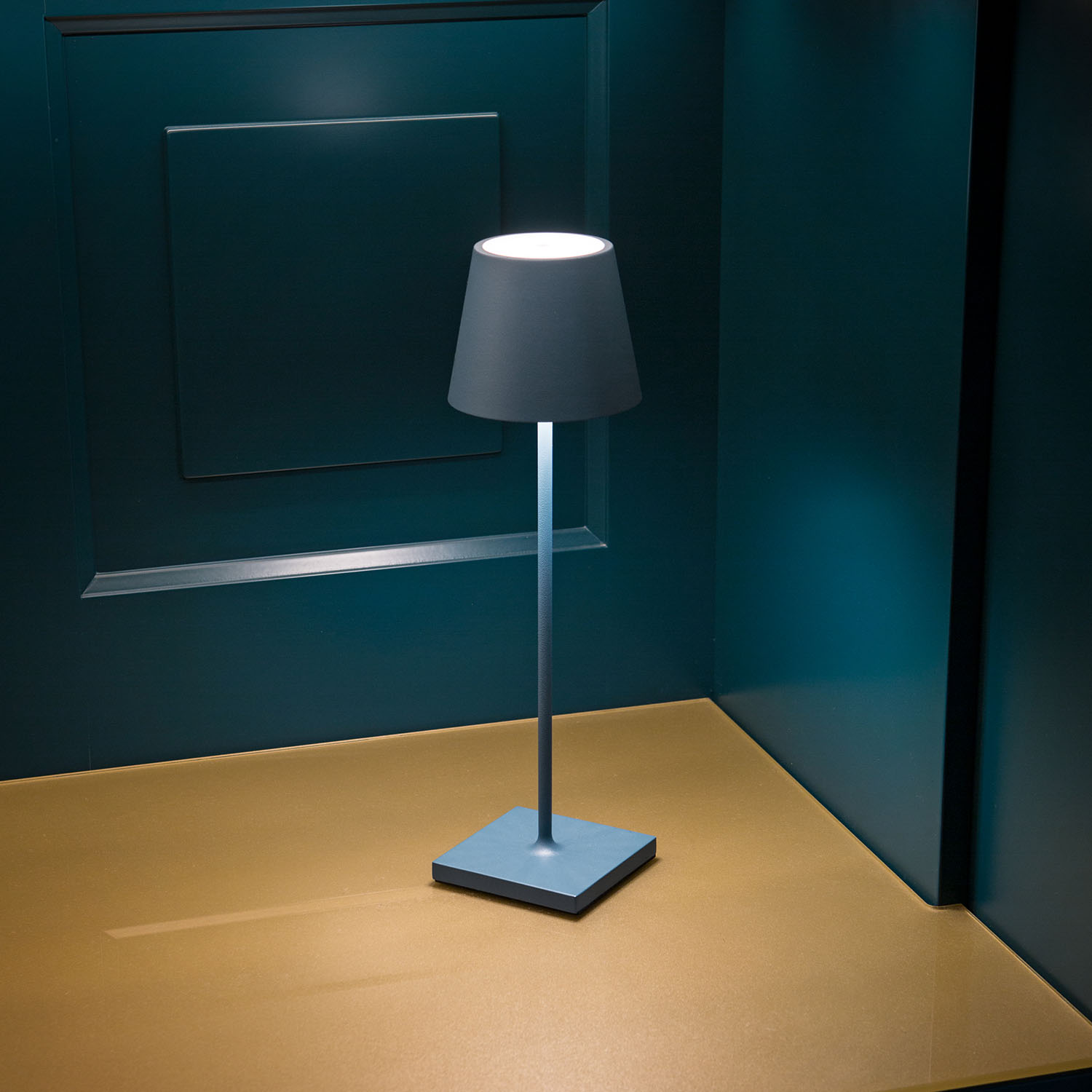 Eine blaue Tischlampe mit Lampenschirm, auf einem Tisch. Die Wand ist blau und der Boden gelb.