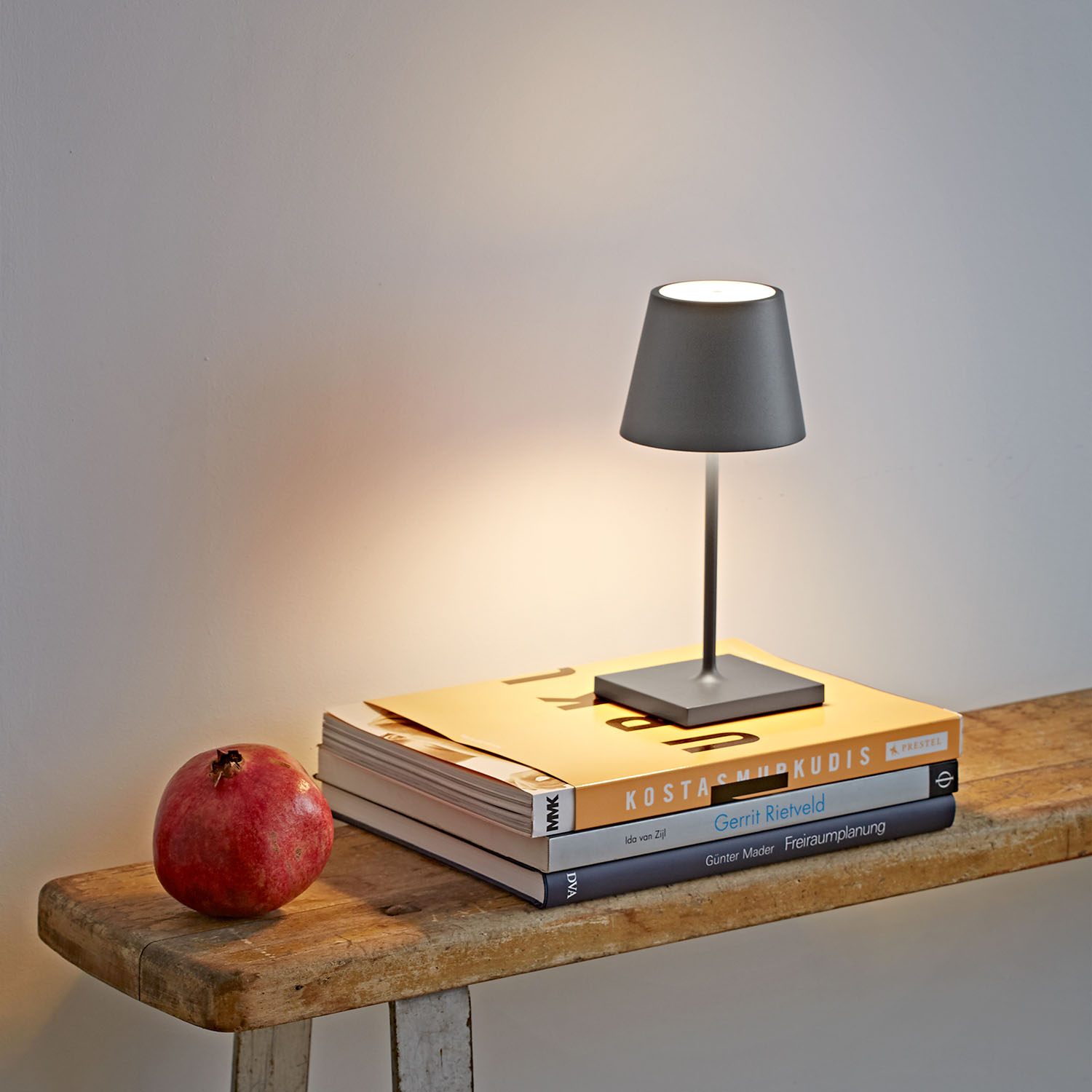 Eine kleine graue Tischlampe auf einem Stapel Bücher und einem roten Granatapfel. Der Hintergrund ist eine Holzbank und eine weiße Wand.