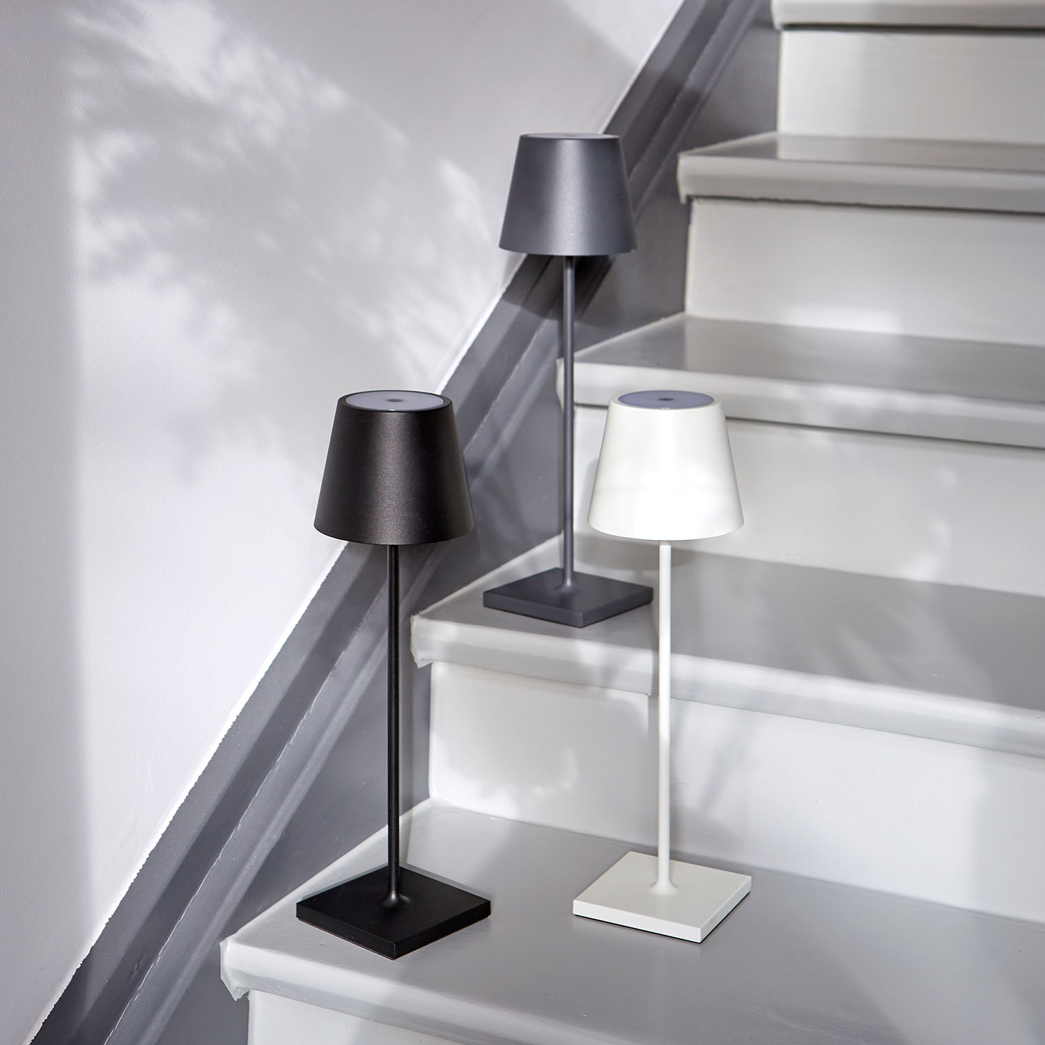 Drei kleine Lampen auf Treppen. Eine ist schwarz, eine grau und eine weiß. Der Hintergrund ist eine Wand.
