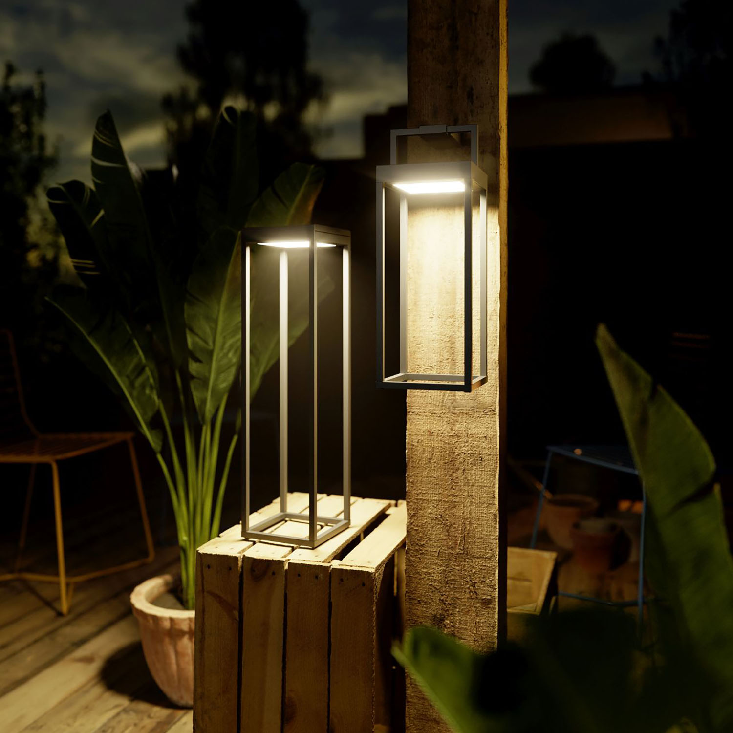 Zwei Außenlampen, eine hochrechteckige, eine quadratische, auf einer Holzkiste, Nachtszene mit Pflanzen und dunklem Himmel.