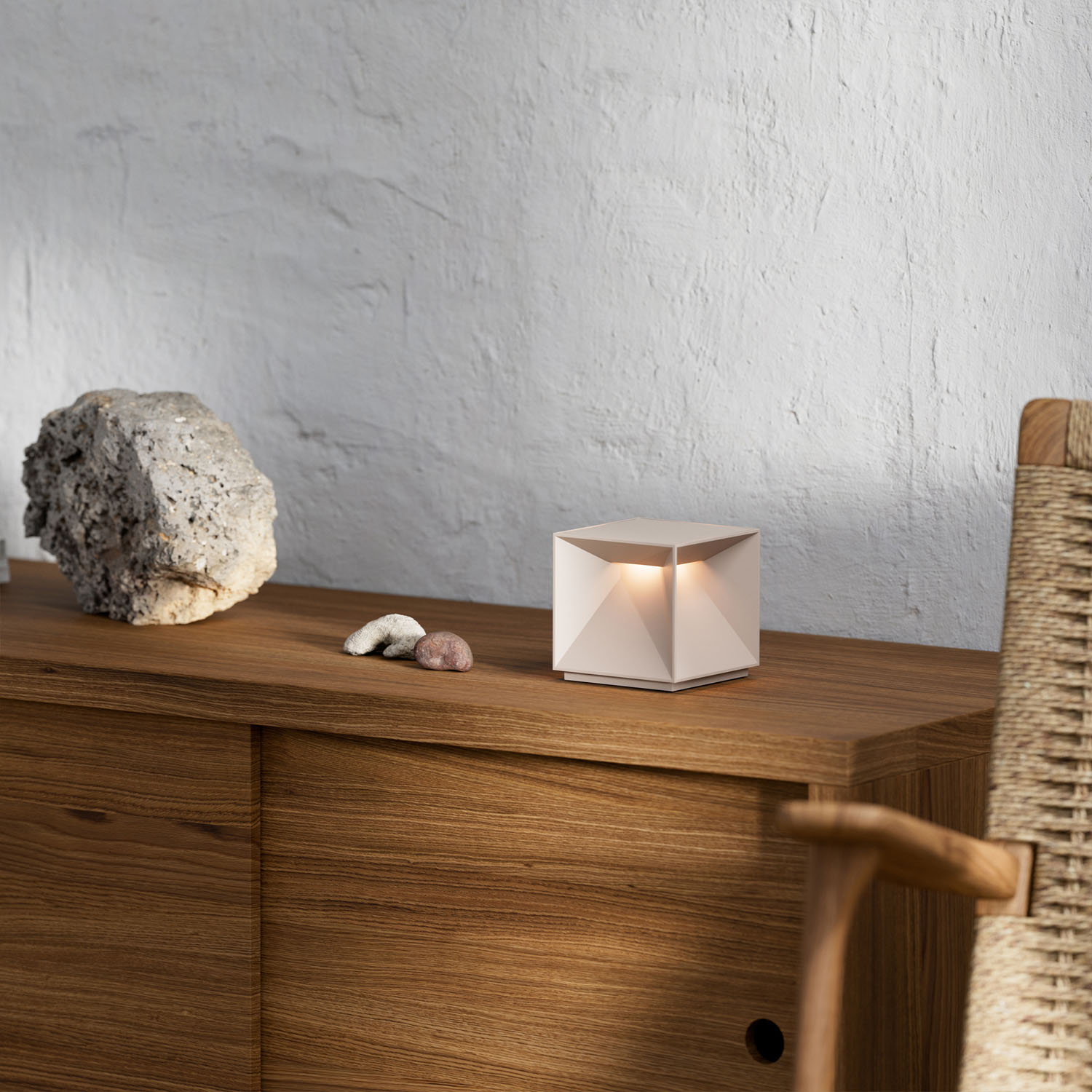 Eine beleuchtete, geometrische Lampe steht auf einem Holzschrank neben einem Stein und einem Stuhl.