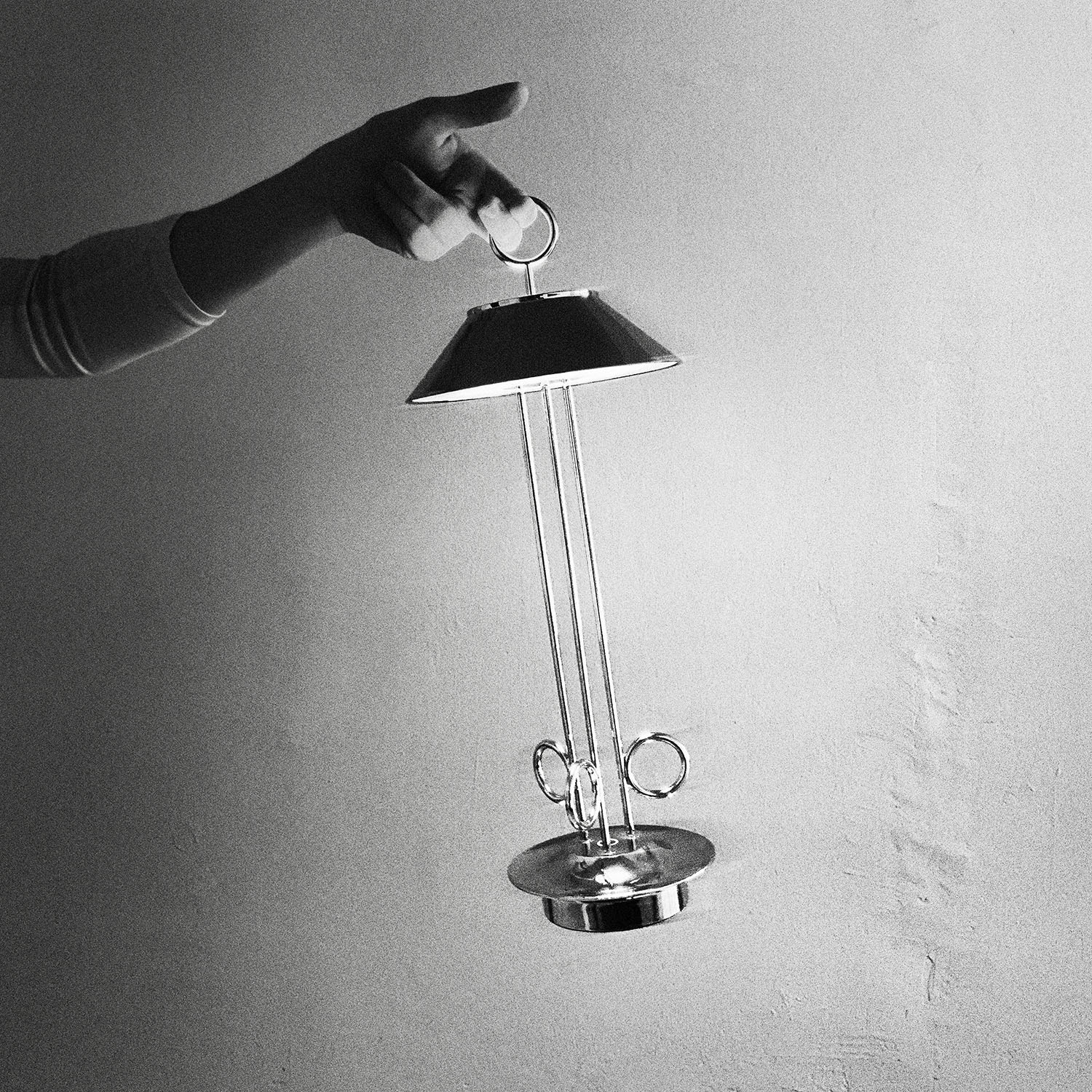 Eine Hand, die eine silberne Lampe mit Sockel hält. Die Lampe hat einen Schirm und drei vertikale Stützen. Schwarz und weiß.